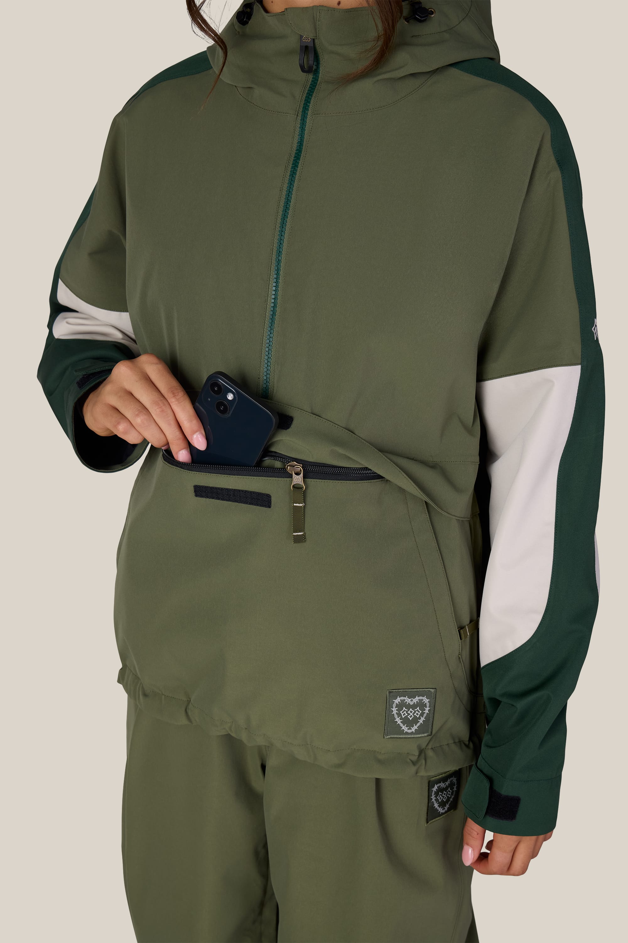 A person wearing the 686 Women’s Outline™ Shell Anorak in green, with matching green pants, places a black smartphone into the front zippered pocket of the jacket. Their head is partially out of frame.