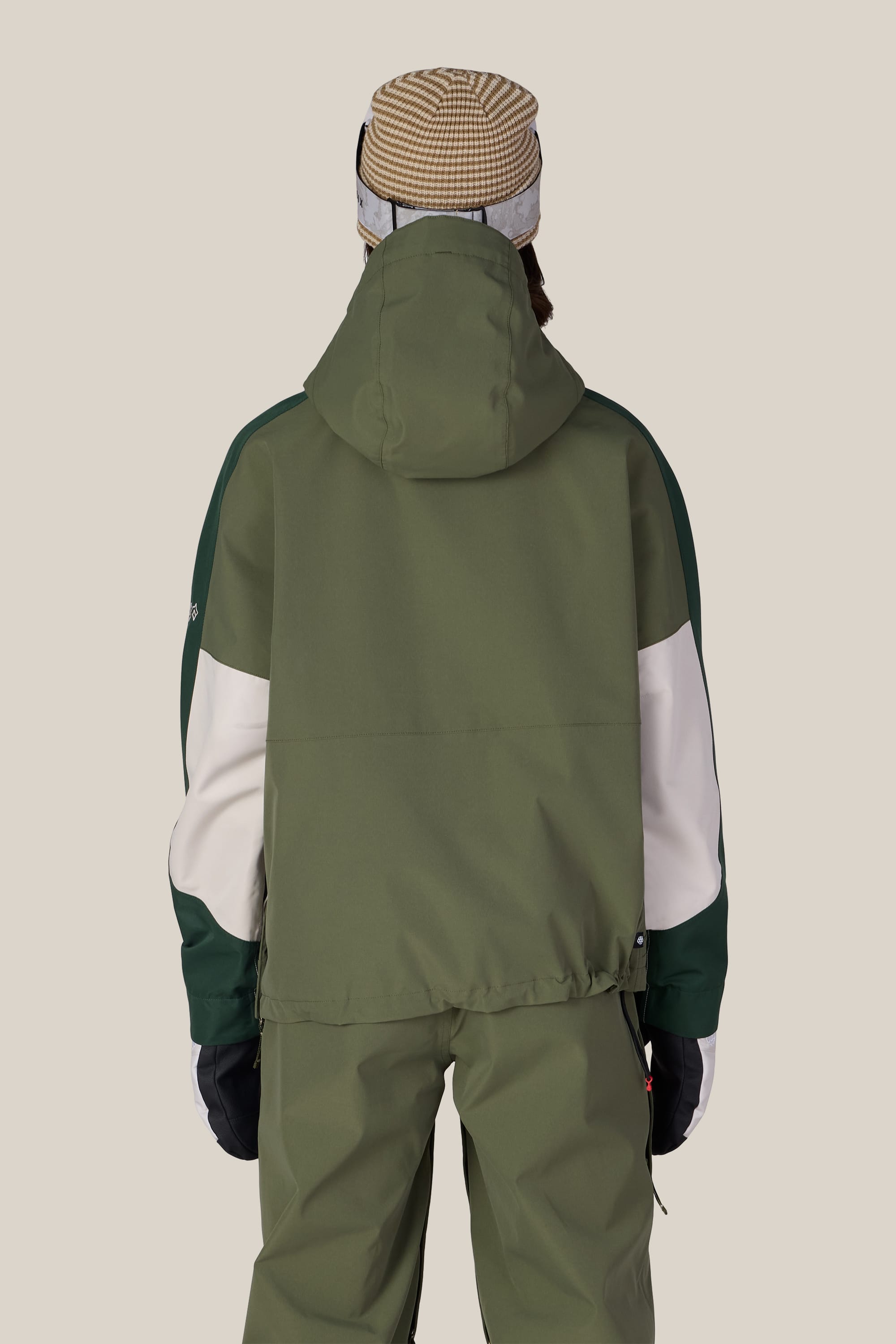 A person is seen from behind against a plain background, wearing the 686 Women’s Outline™ Shell Anorak in olive green with matching pants, black gloves, and a brown striped beanie.