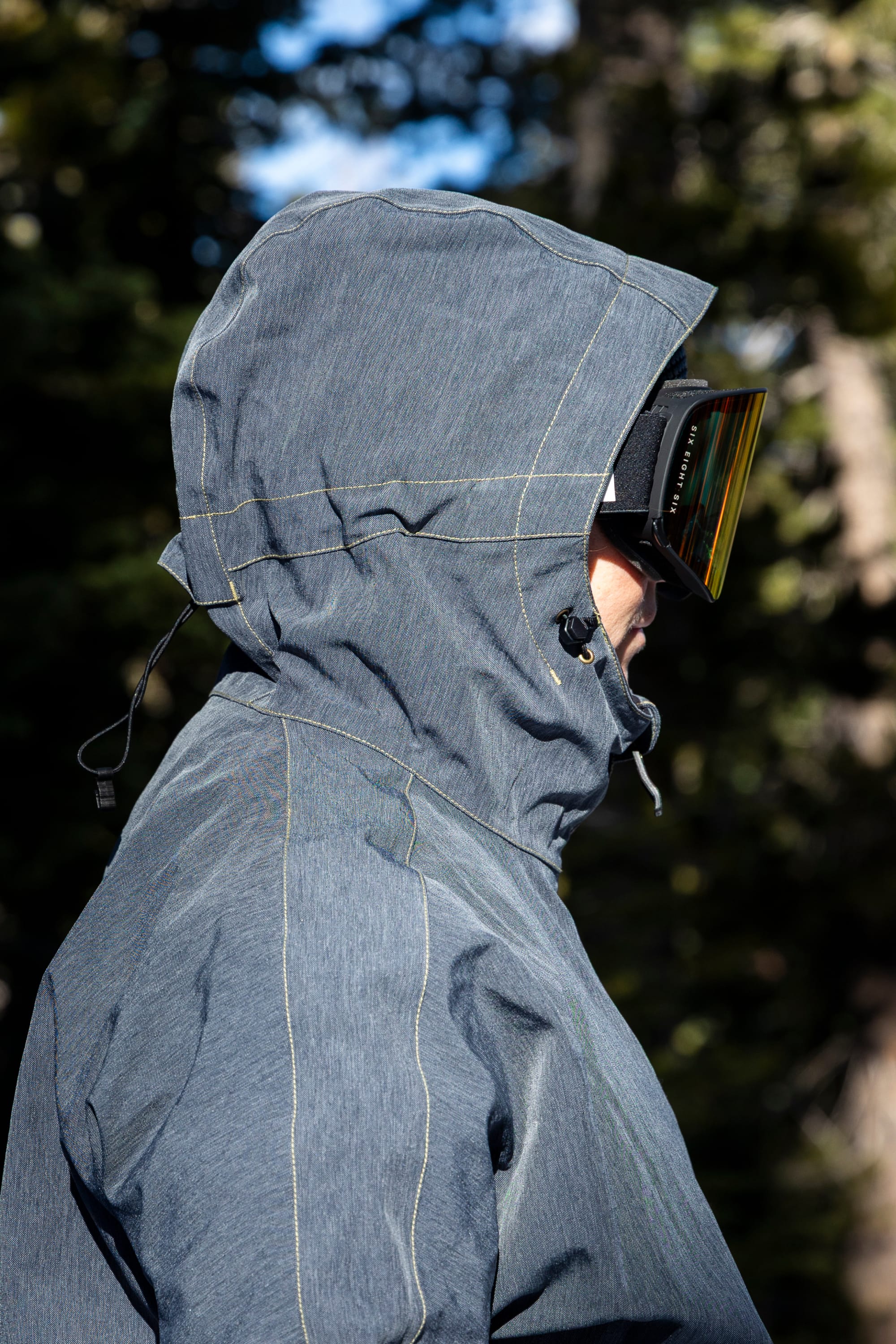 A person in profile wearing the 686 Women’s Outline™ Shell Anorak and ski goggles stands outdoors with sunlight and blurred trees in the background.