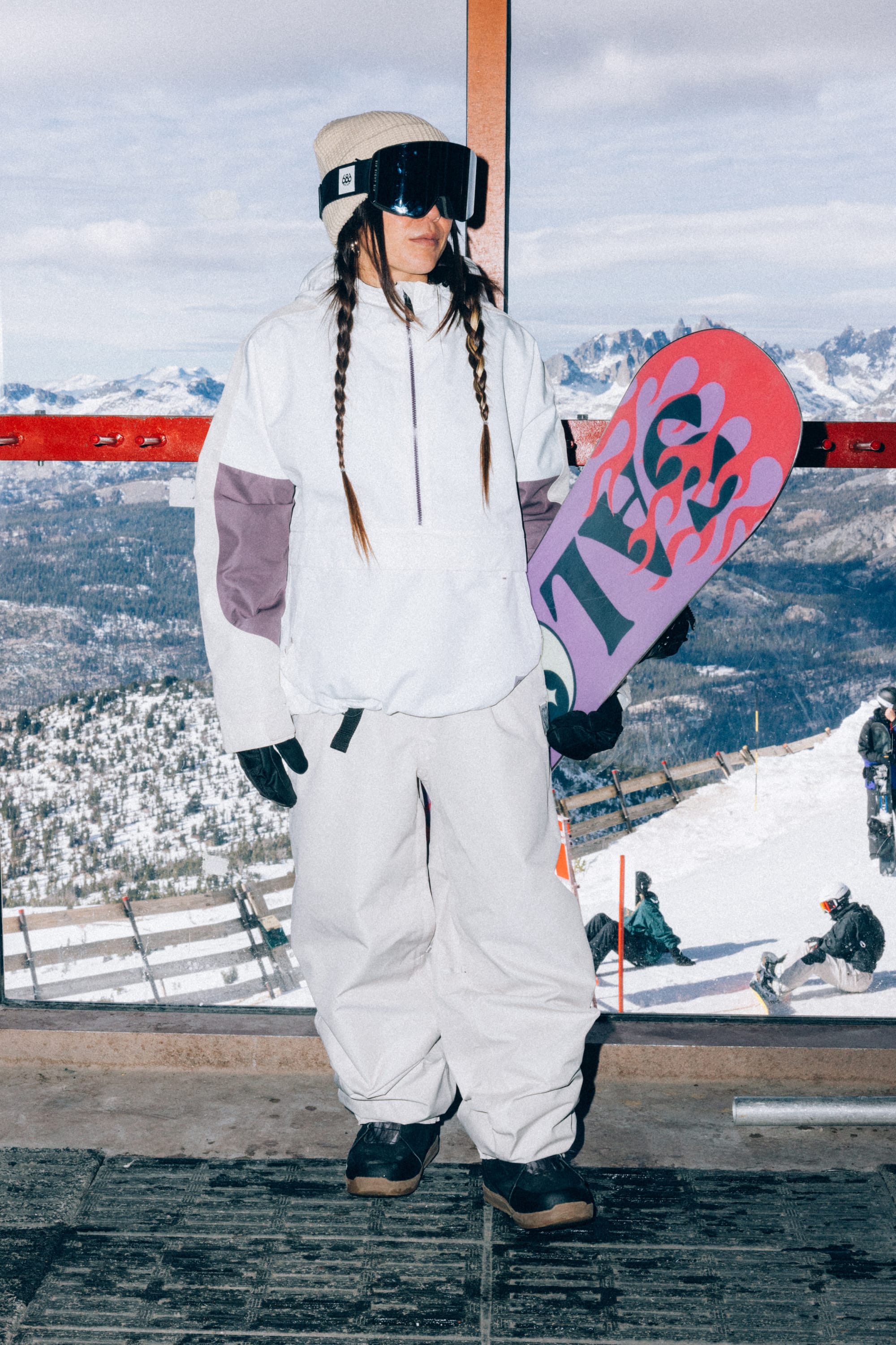 Wearing the 686 Women’s Outline™ Shell Anorak, a snowboarder stands indoors in white winter gear and goggles, holding a pink and purple snowboard. Snowy mountains and other people on a ski slope are visible through the window behind them.