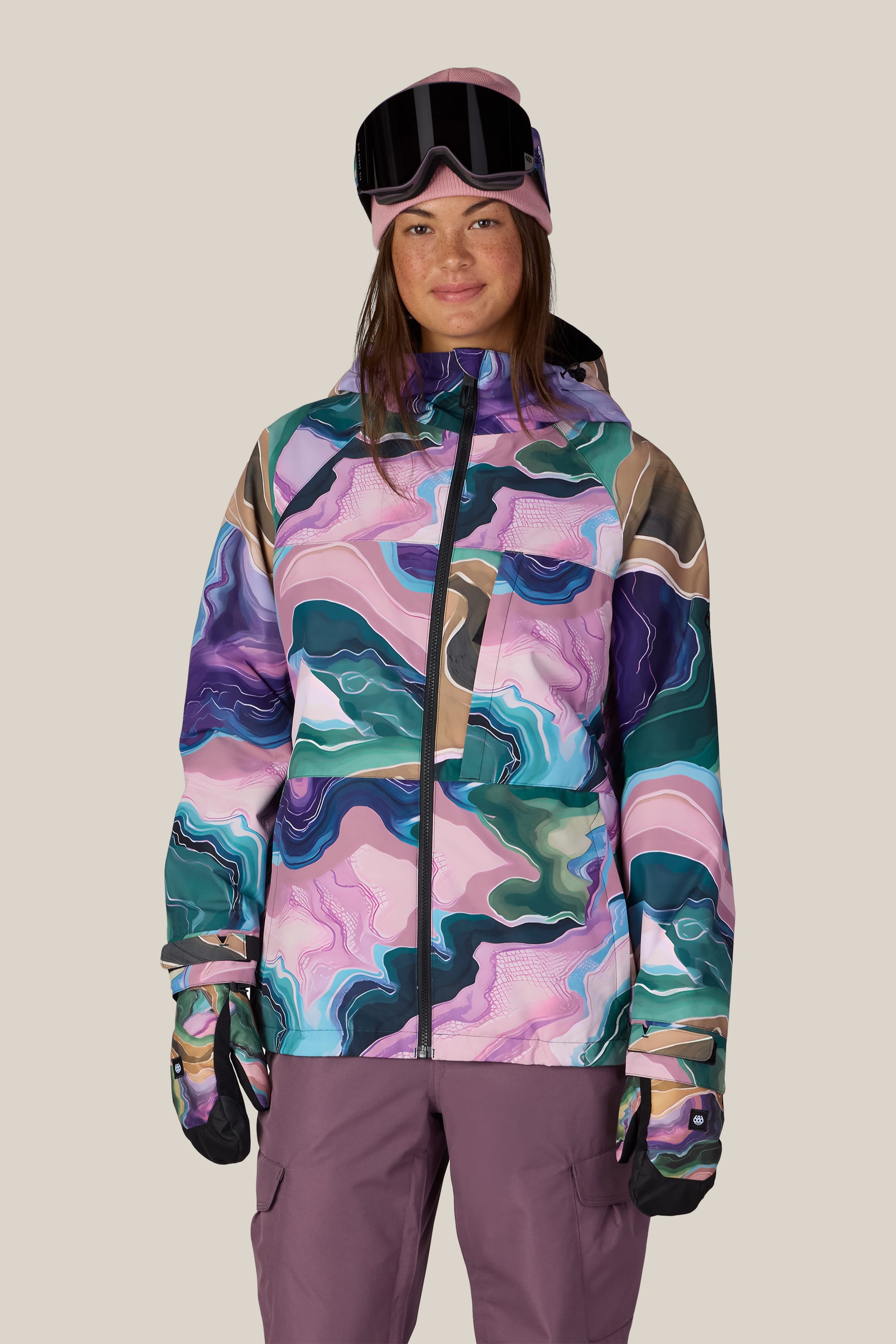 A woman stands facing the camera, smiling slightly, against a plain light background. She is wearing the 686 Women’s Athena™ Insulated Jacket by 686, which features a colorful, abstract pattern, along with purple snow pants, gloves, and goggles.