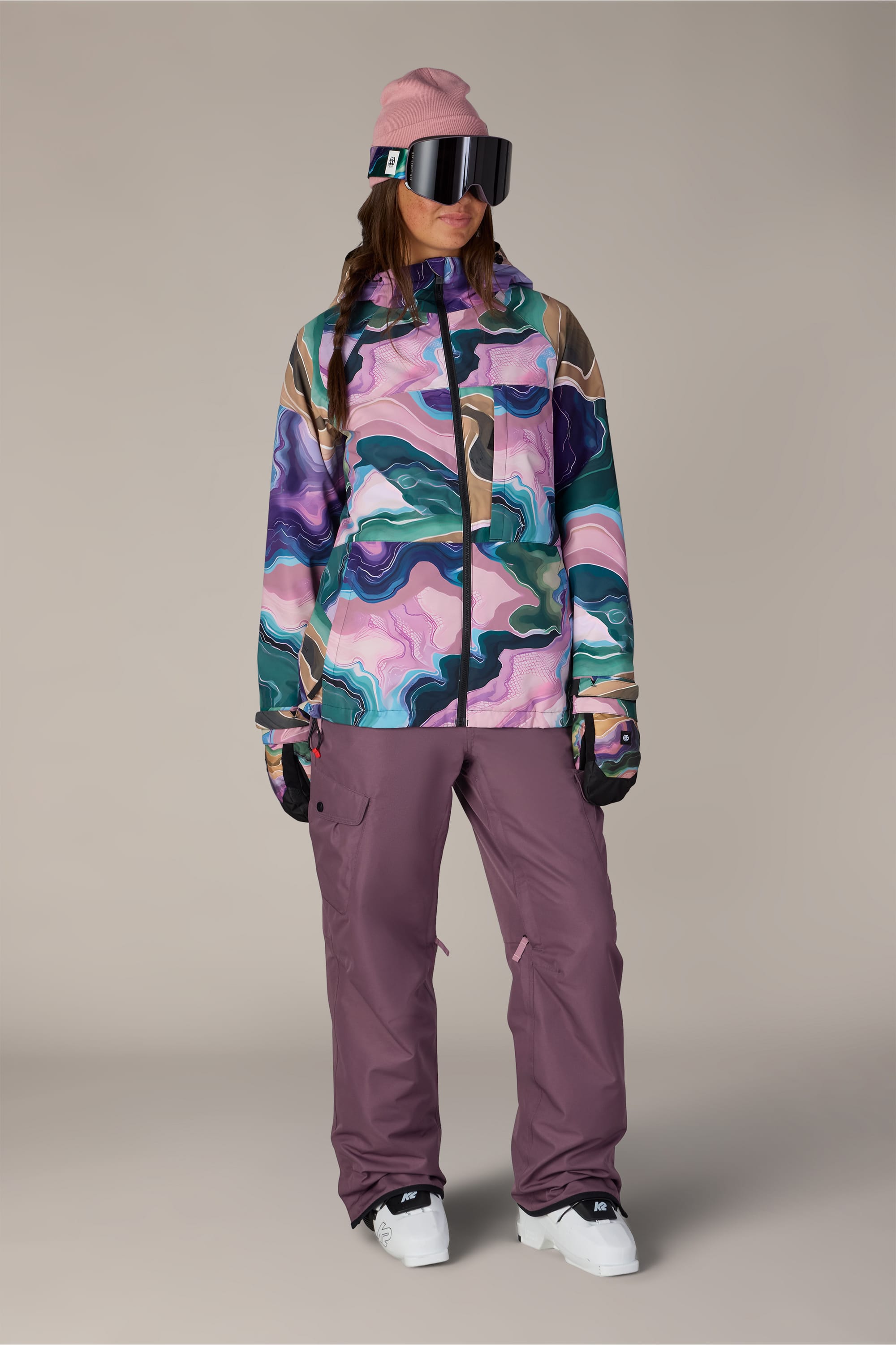 A person wearing the 686 Women’s Athena™ Insulated Jacket by 686, styled with mauve snow pants, black gloves, a pink beanie, large ski goggles, and white snow boots, standing against a plain background.