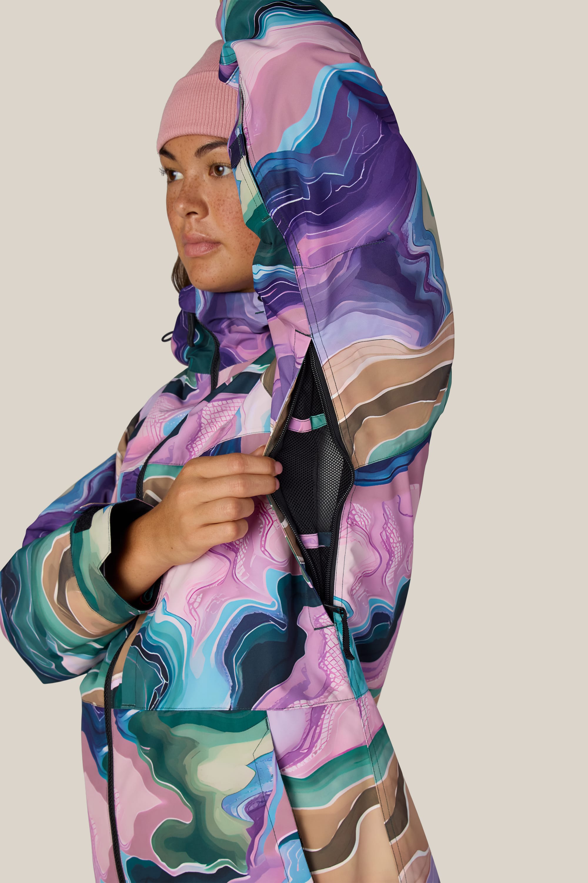 A person wearing the 686 Women’s Athena™ Insulated Jacket and a pink beanie opens a black mesh-lined zipper under their arm, demonstrating the jacket’s ventilation feature against a plain beige background.
