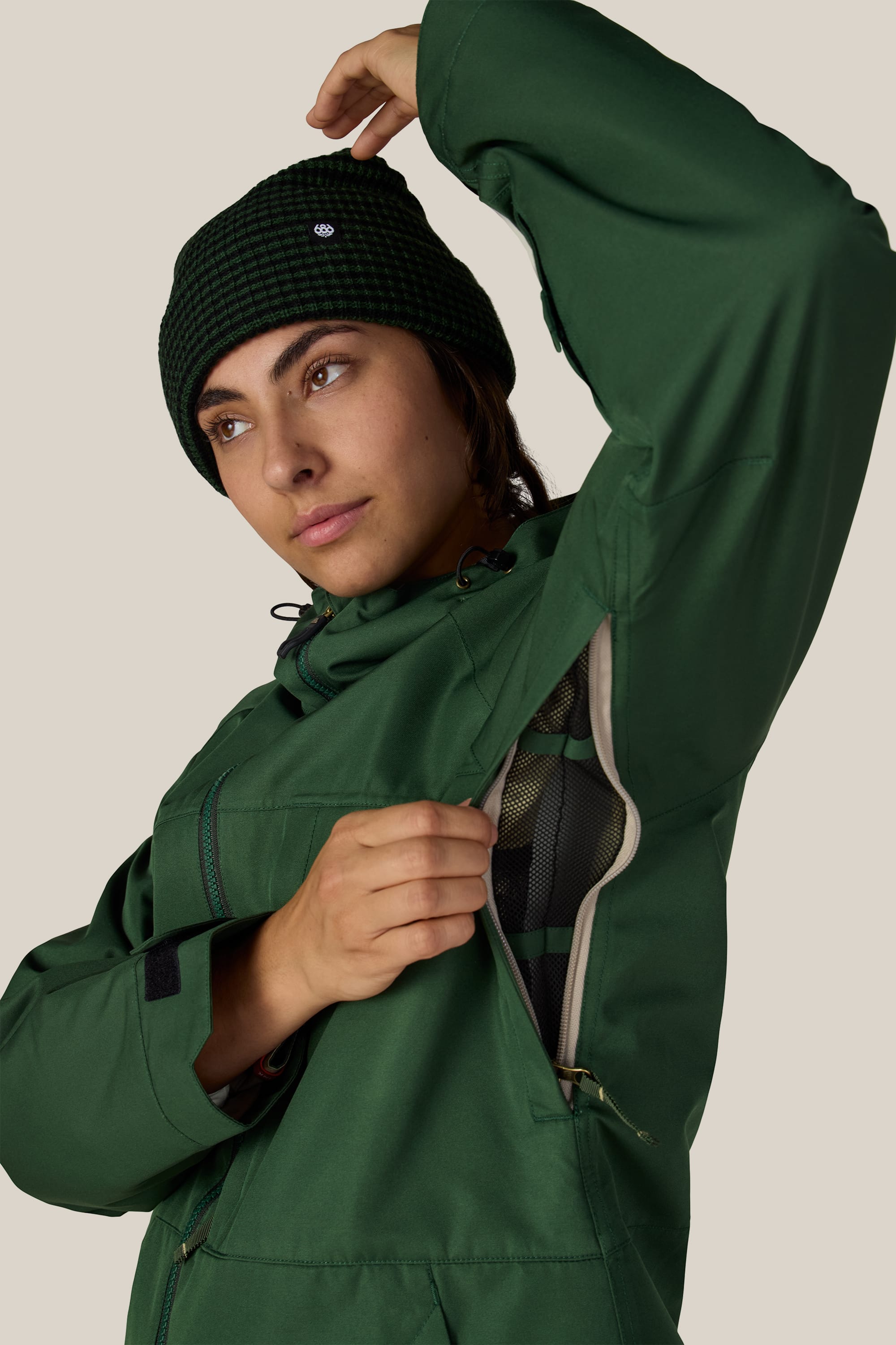 A person wearing the 686 Women’s Athena™ Insulated Jacket in green and a dark beanie unzips the underarm vent to reveal the inner lining, set against a neutral background.