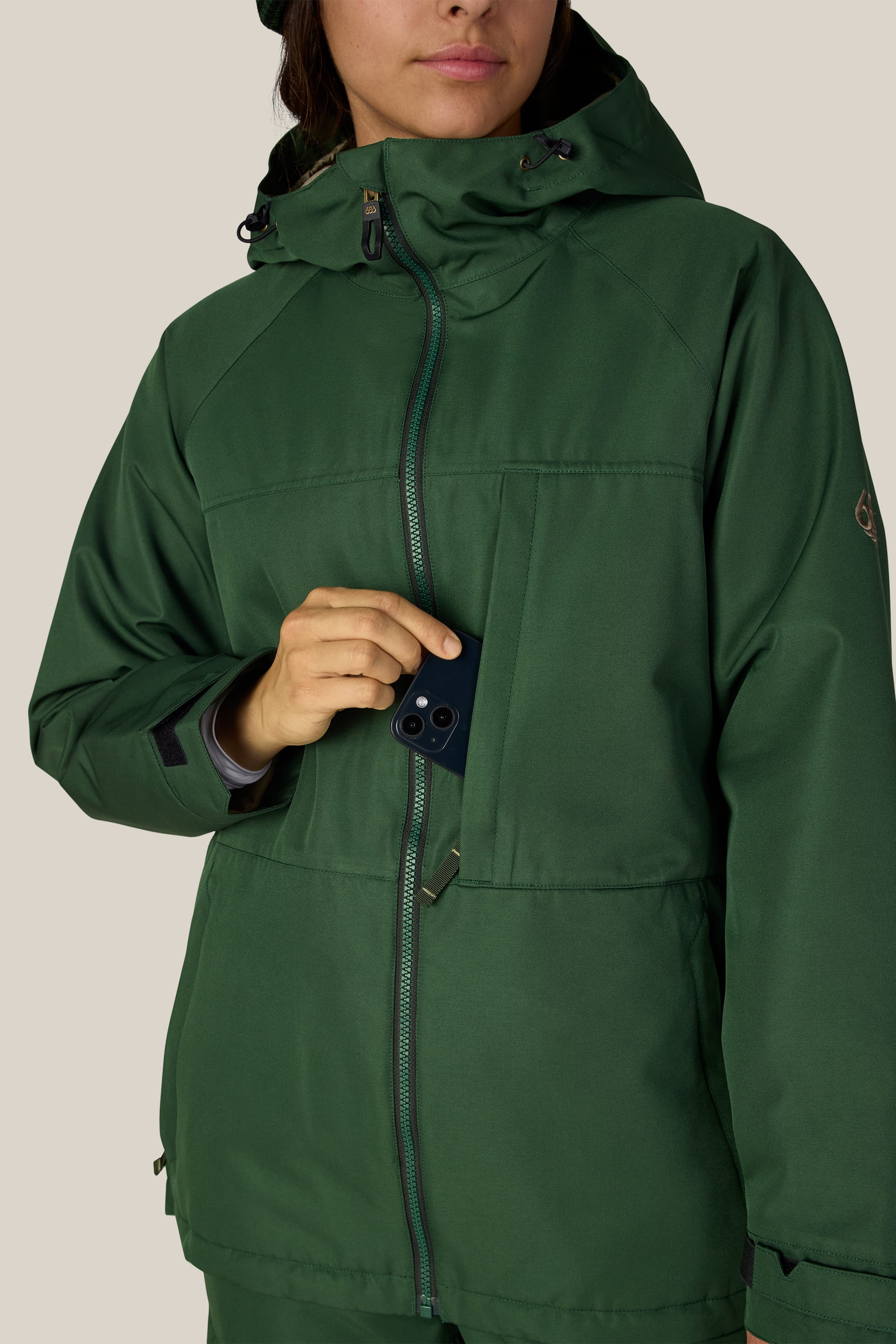 A person wearing the 686 Women’s Athena™ Insulated Jacket places a black smartphone into a chest pocket, with only their lower face and upper body visible against a plain background.