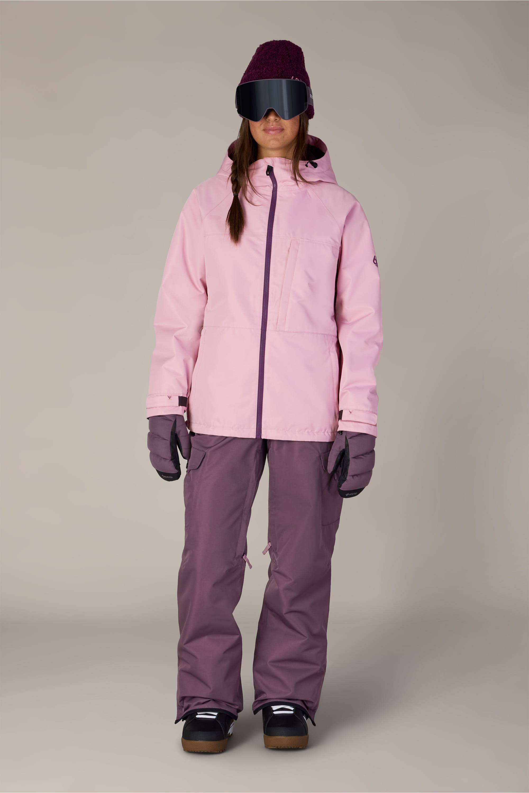 A person is wearing the 686 Women’s Athena™ Insulated Jacket in light pink, paired with purple snow pants, gloves, dark snow boots, a maroon beanie, and black ski goggles while standing facing forward against a plain background.