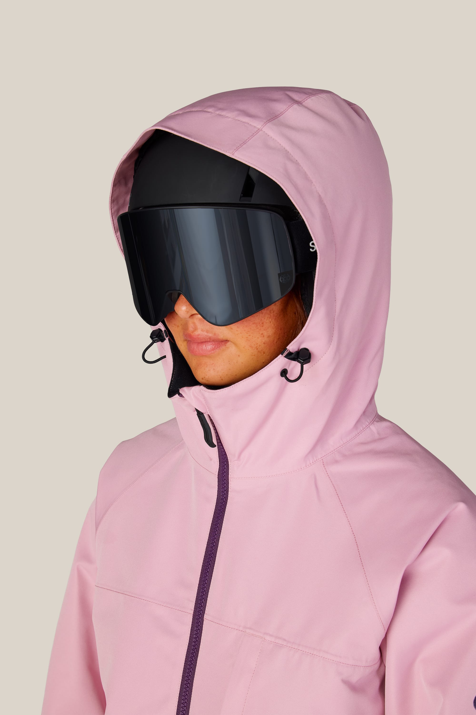 A person dressed in the 686 Women’s Athena™ Insulated Jacket in light pink, along with black ski goggles and a black helmet, stands slightly turned to the side against a plain beige background.