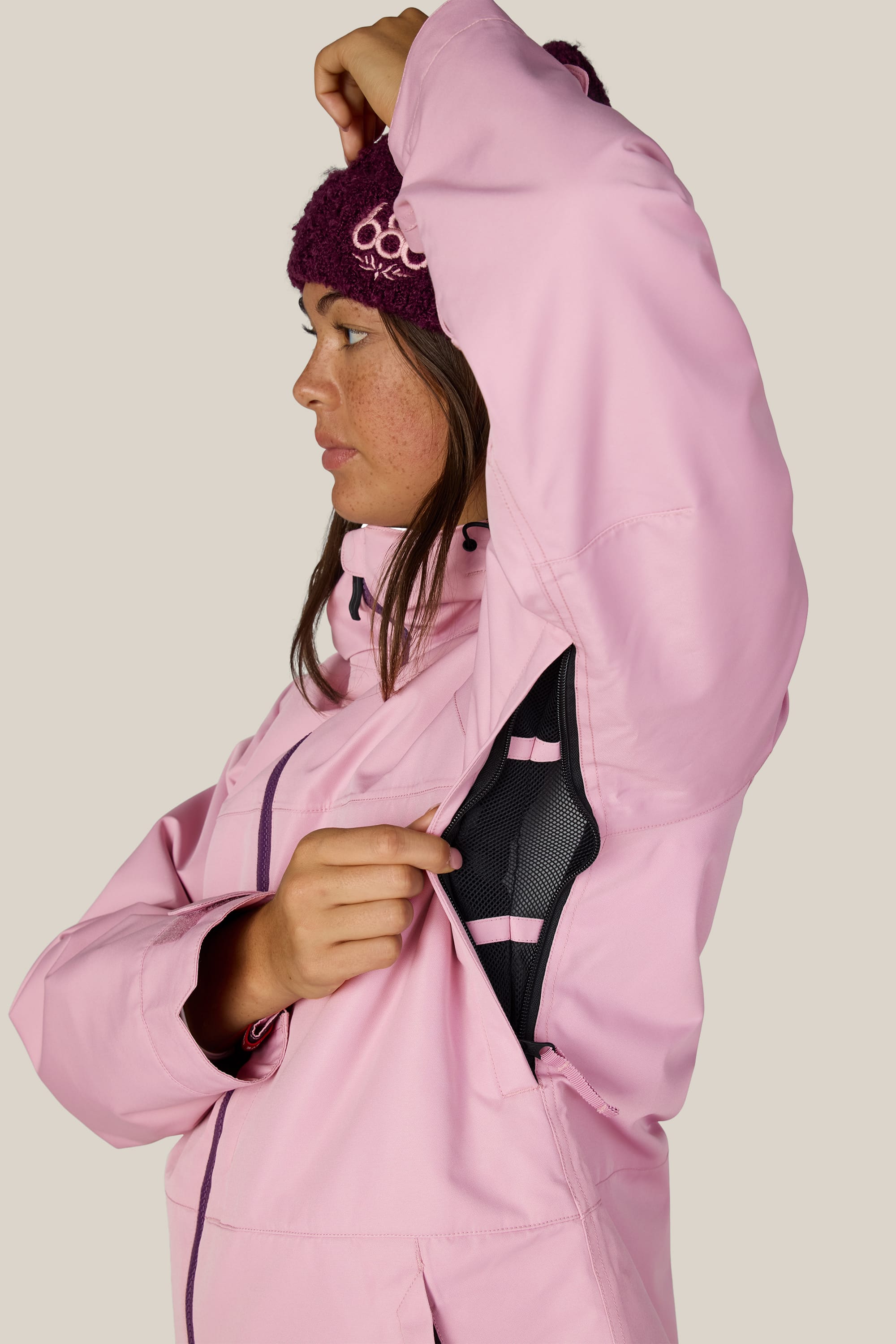 Wearing the 686 Women’s Athena™ Insulated Jacket, a person in a pink jacket and maroon beanie lifts their arm to open a zippered underarm vent, revealing black mesh inside.