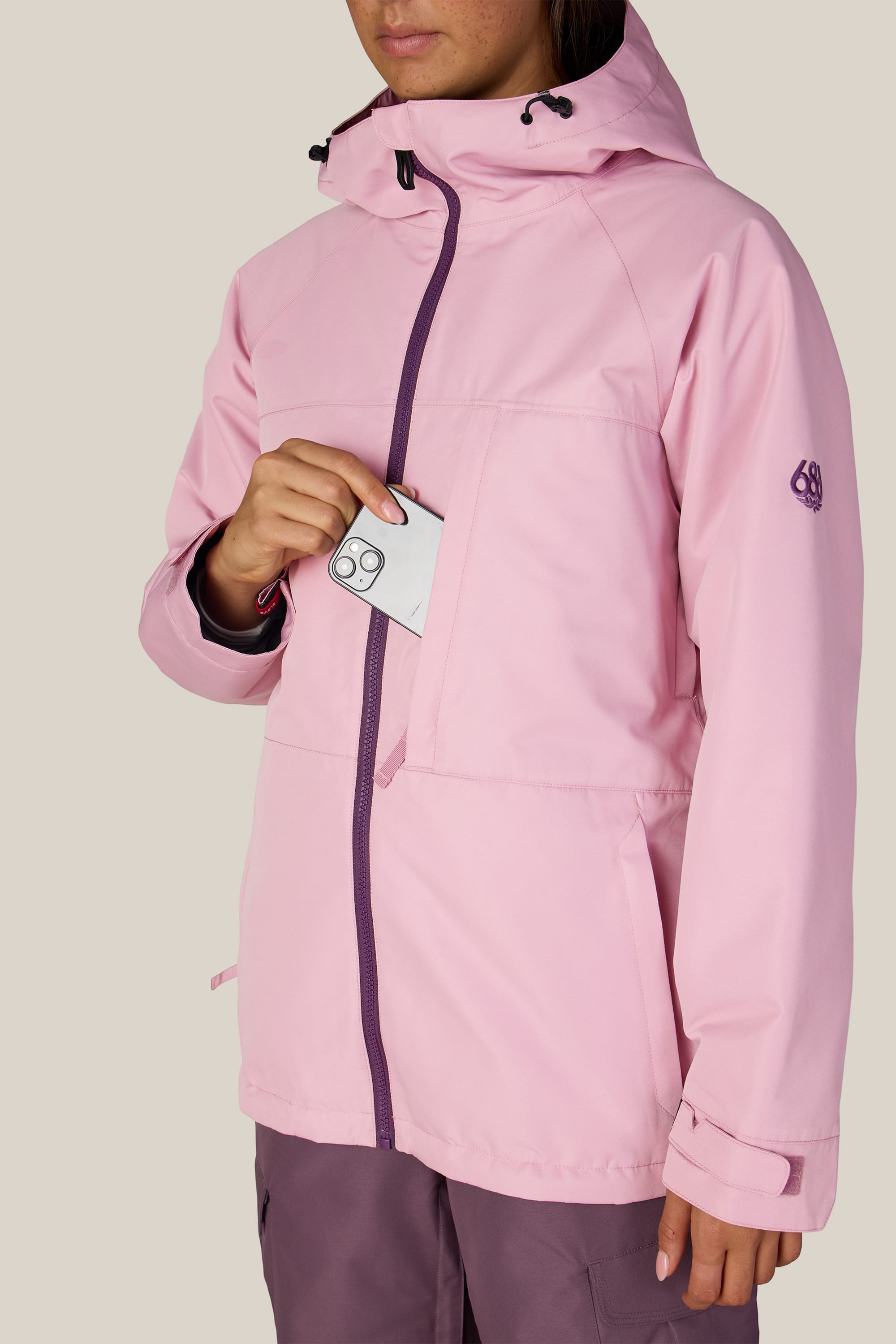 A person wearing the 686 Women’s Athena™ Insulated Jacket in light pink places a card with the number 69 into the chest pocket. Their face is partially visible against a plain beige background.