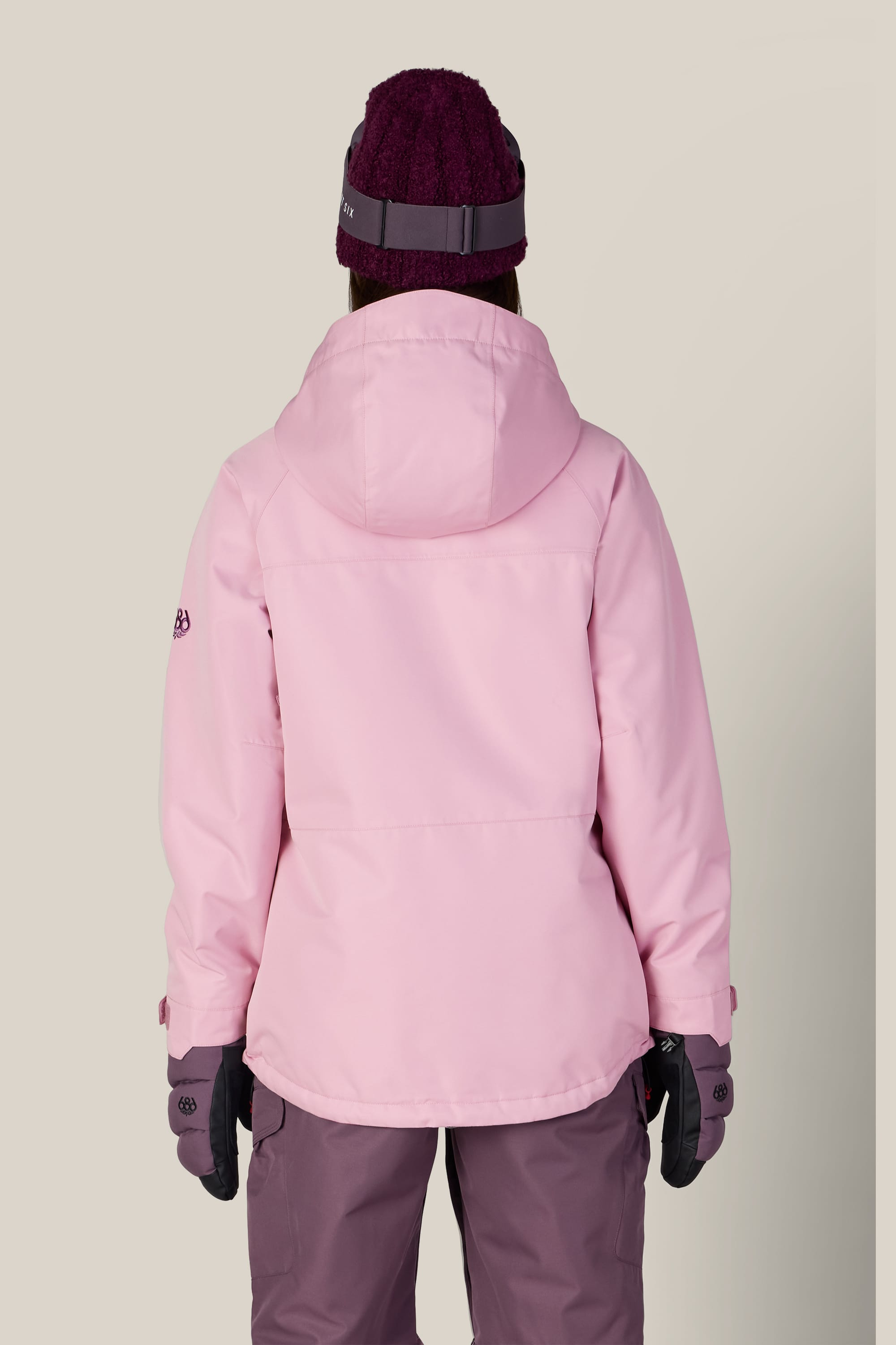 A person is standing with their back to the camera against a plain light background, wearing the 686 Women’s Athena™ Insulated Jacket in pink by 686, along with purple gloves, a maroon knit hat, and mauve pants.