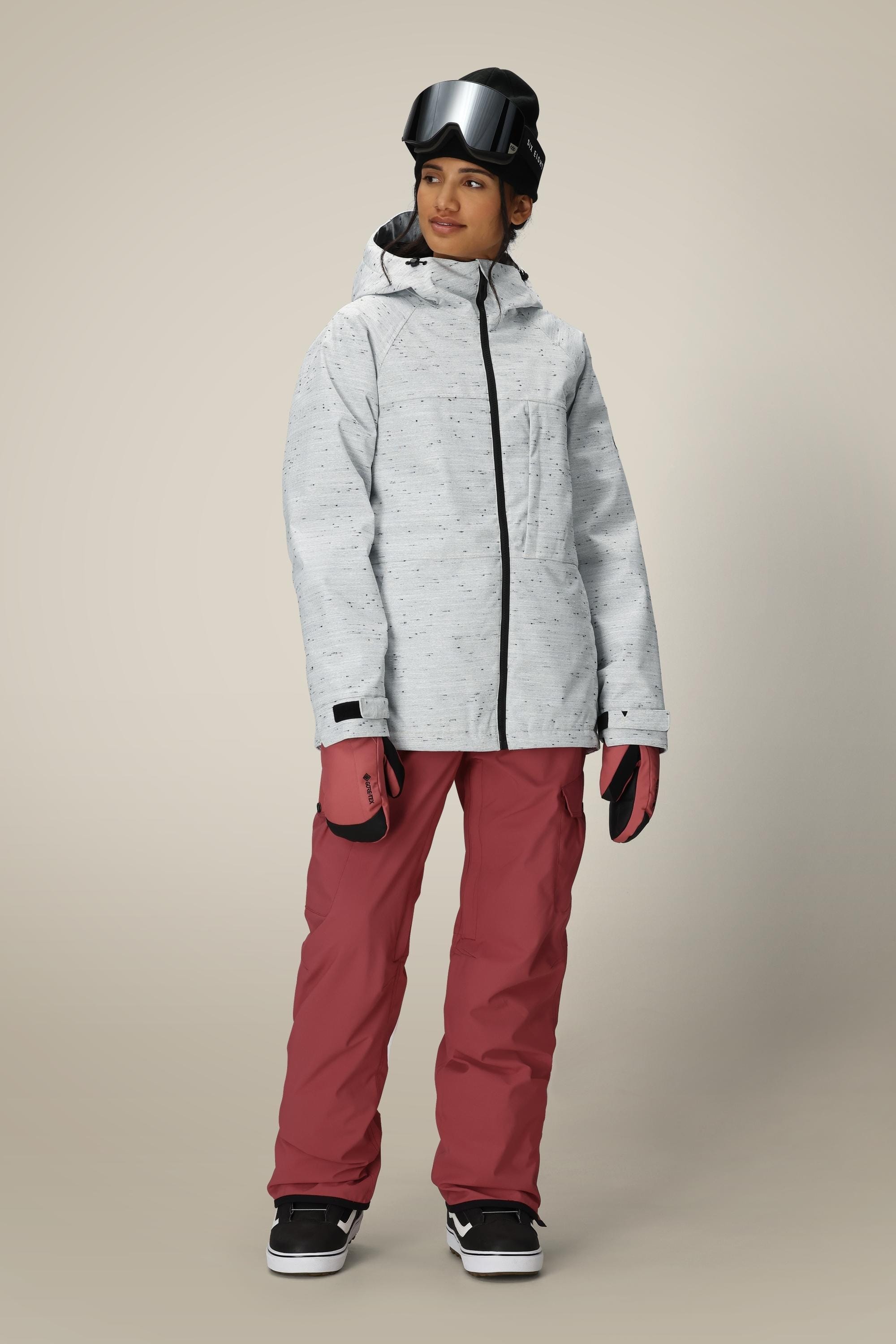 A person wears the 686 Women’s Athena™ Insulated Jacket in light gray, paired with mauve snow pants, black gloves, and ski goggles, standing against a neutral background.