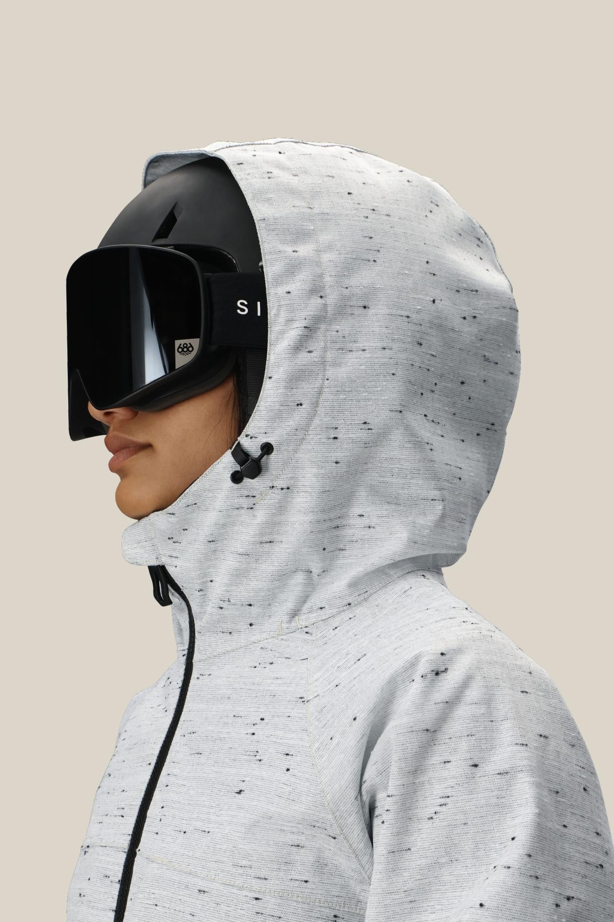 A person wearing the 686 Women’s Athena™ Insulated Jacket in a speckled light gray, paired with a black ski helmet and large black ski goggles, is shown in profile against a plain beige background.