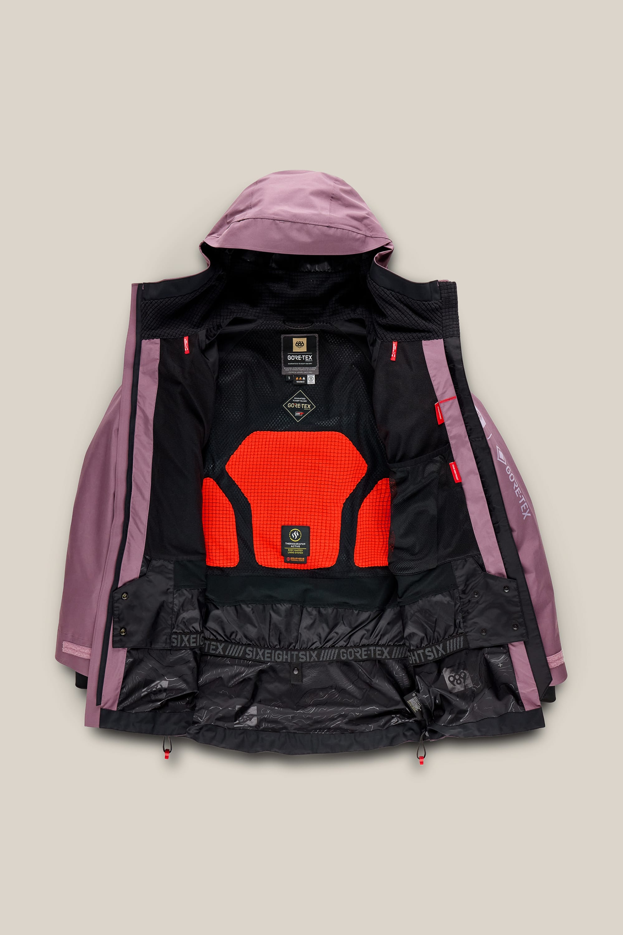 A 686 Women’s GORE-TEX® Skyline Thermagraph® Jacket in mauve is laid flat, unzipped to reveal a black and orange mesh inner lining, with 686 branding on the bottom hem, displayed against a neutral beige background.