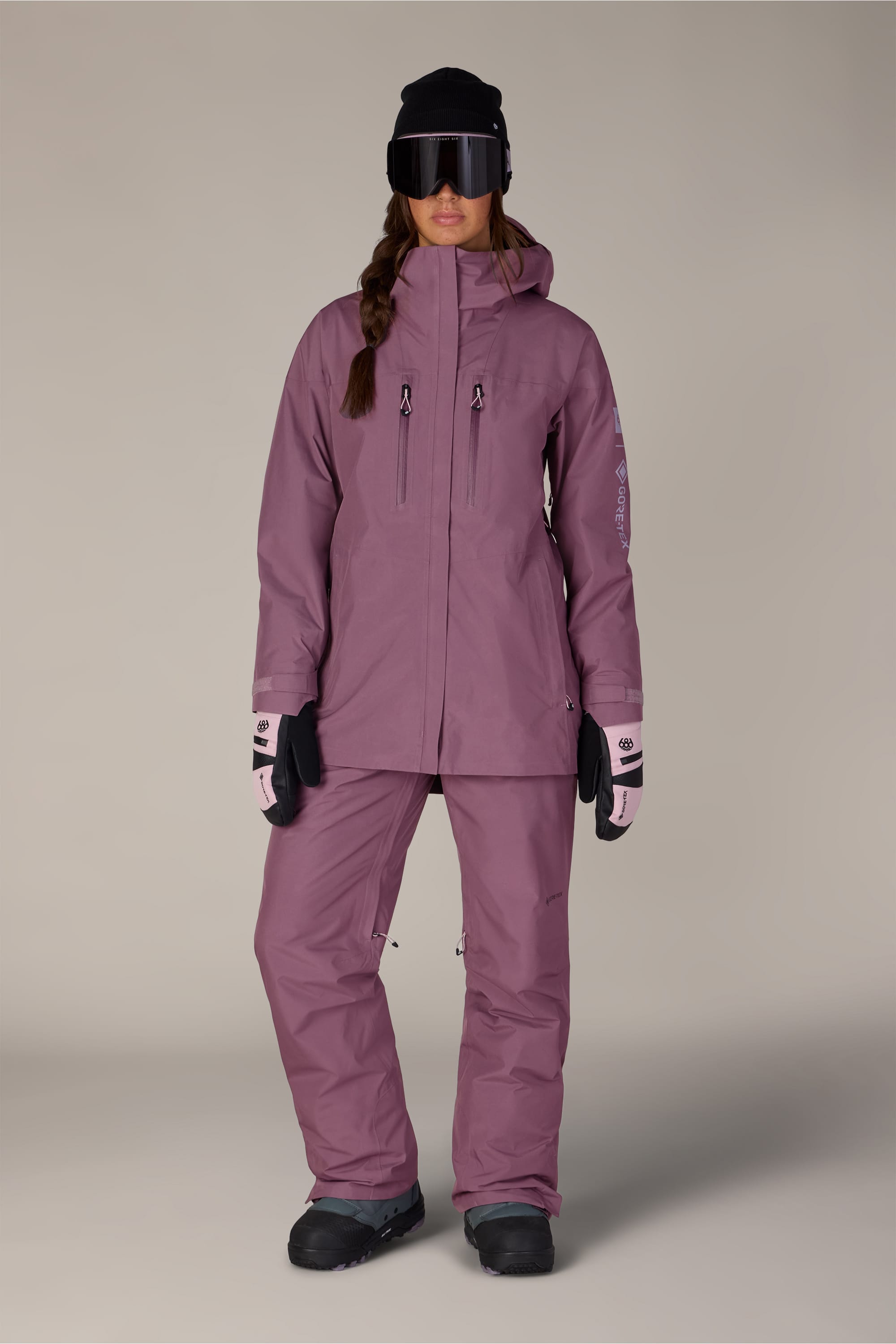 A person stands facing forward, wearing the 686 Women’s GORE-TEX® Skyline Thermagraph® Jacket in mauve, paired with matching pants, black boots, light pink gloves, black ski goggles, and a black beanie against a plain background.