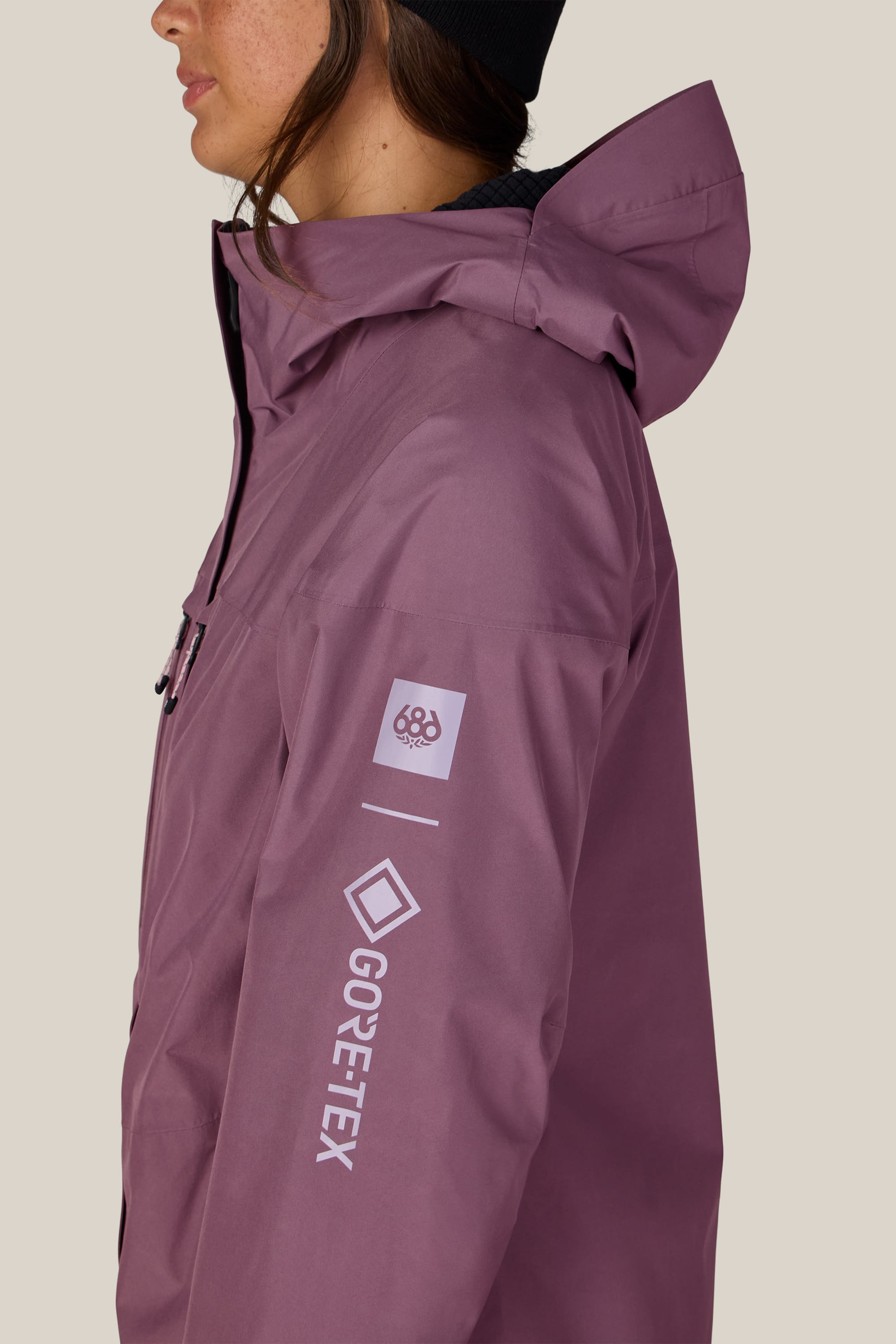 A person is shown from the upper body, partially facing left, wearing a mauve 686 Women’s GORE-TEX® Skyline Thermagraph® Jacket with hood and black beanie. The 686 logo is visible on the sleeve against a plain background.