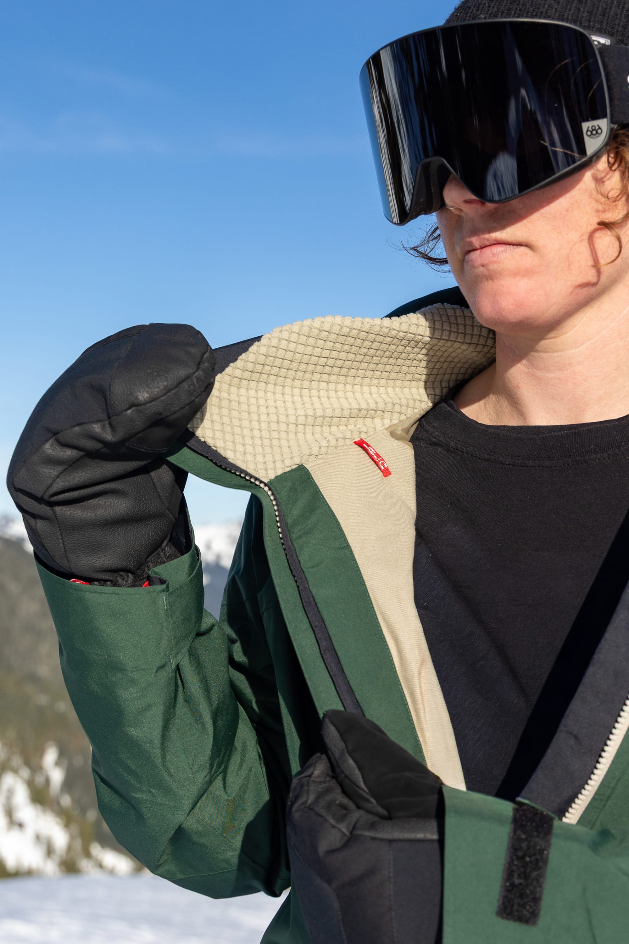 A person wearing black ski gloves, large black ski goggles, and the 686 Women’s GORE-TEX® Skyline Thermagraph® Jacket by 686 with a tan lining stands outdoors in a snowy mountain area under a clear blue sky.