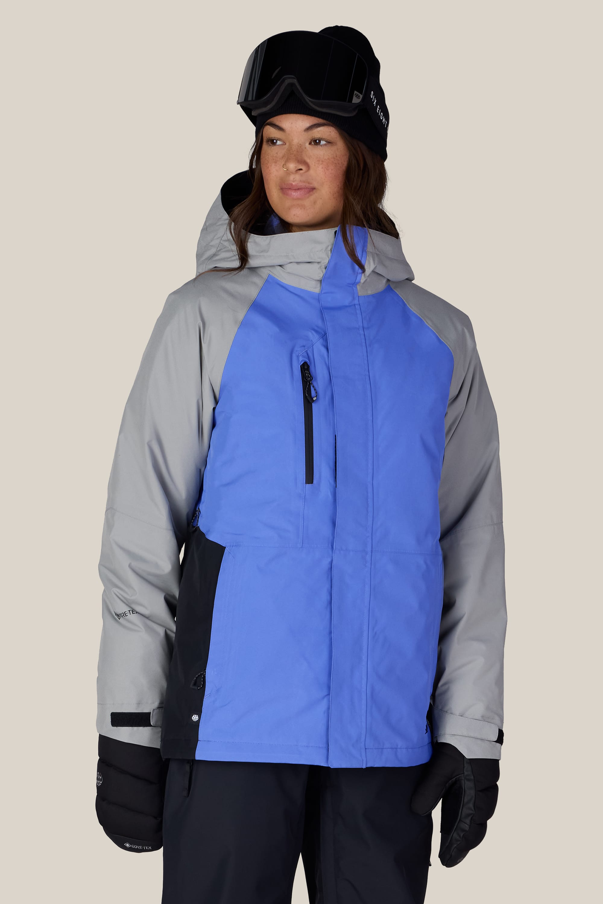 Wearing the 686 Women’s GORE-TEX® Willow™ Insulated Jacket in blue and gray, along with black winter gloves, black ski goggles, and a black beanie, this person stands against a plain light background.