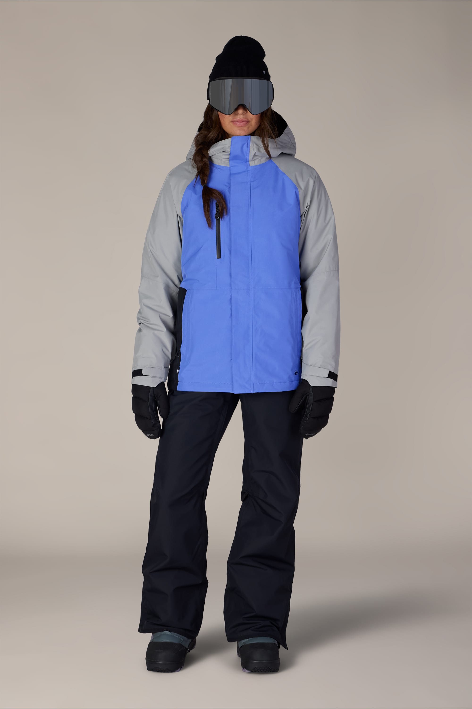 A person is wearing the 686 Women’s GORE-TEX® Willow™ Insulated Jacket by 686 in blue and gray, paired with black snow pants, gloves, boots, a black beanie, and large ski goggles while standing against a plain background.