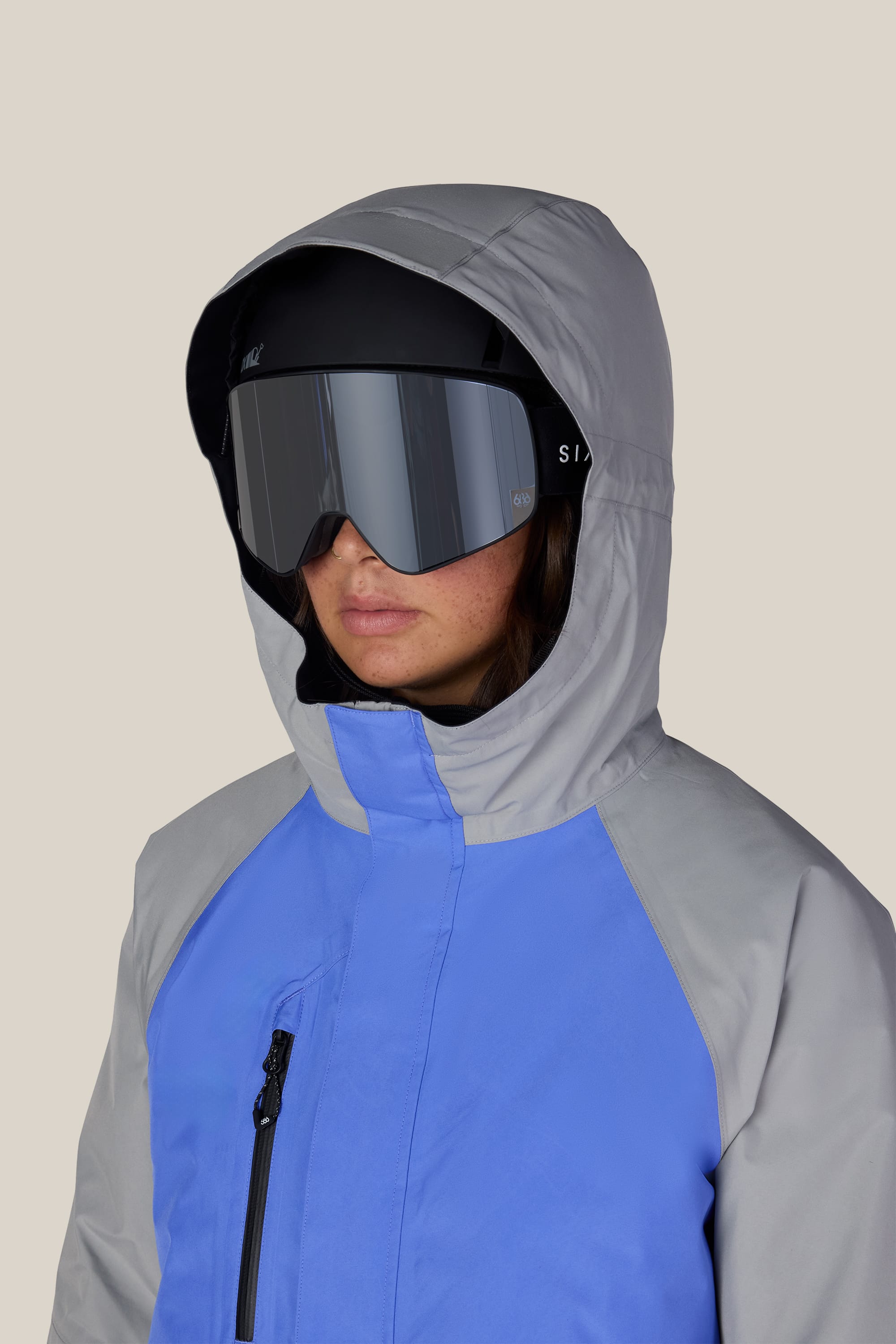 A person wearing the 686 Women’s GORE-TEX® Willow™ Insulated Jacket in gray and blue, along with large black ski goggles, faces slightly left against a plain light background.
