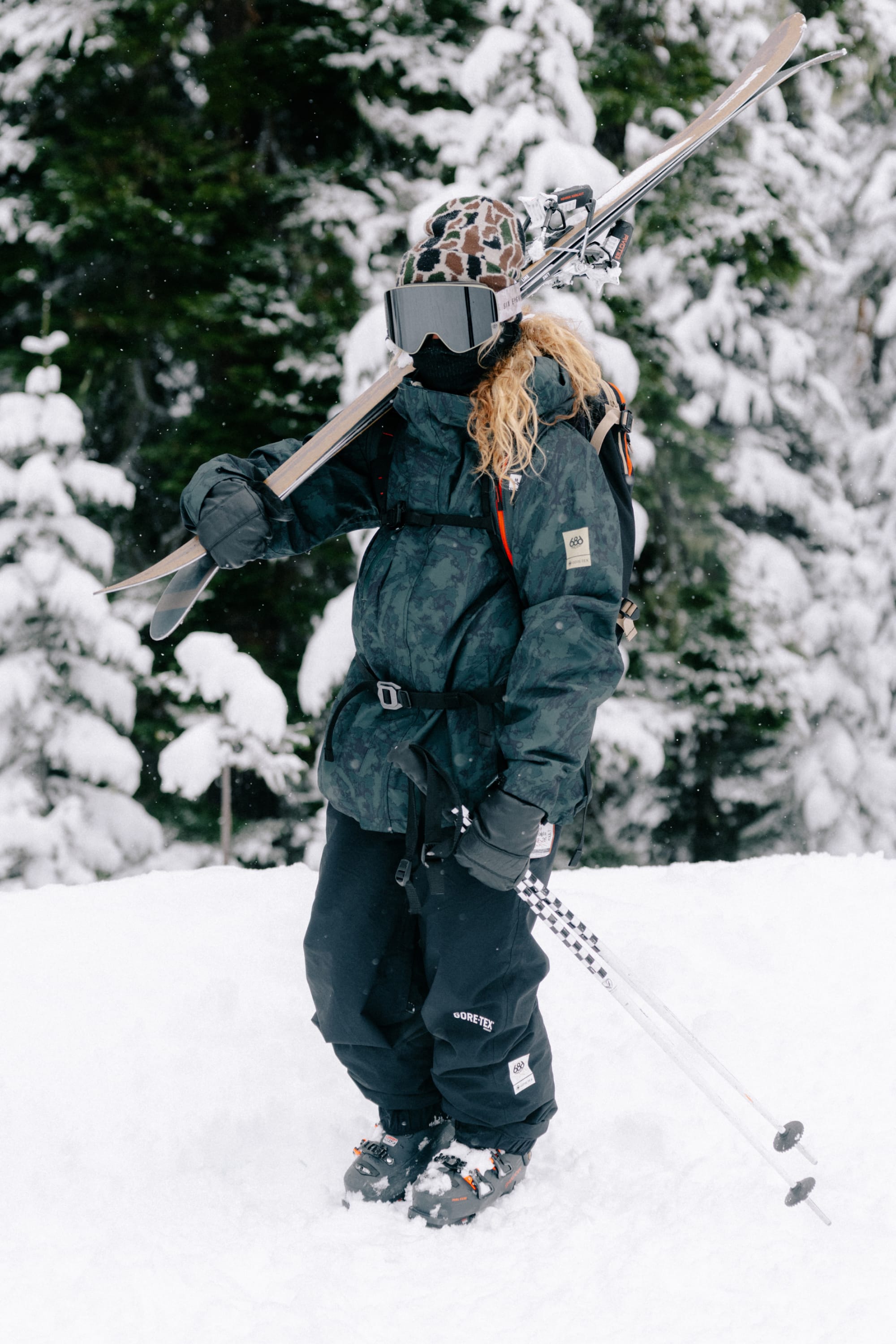 Wearing the 686 Women’s GORE-TEX® Willow™ Insulated Jacket, a skier stands on snowy ground with skis over her shoulder and ski poles in hand, framed by snow-covered trees.