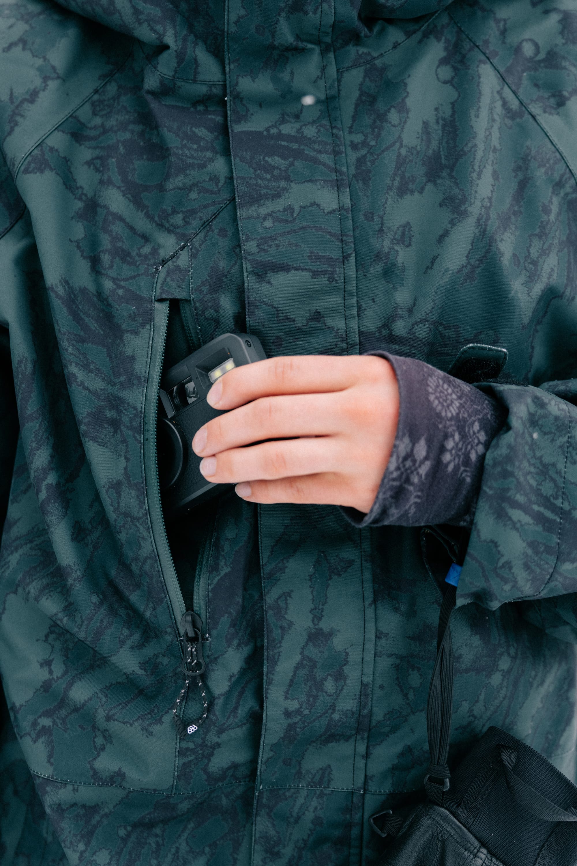 A person wearing the 686 Women’s GORE-TEX® Willow™ Insulated Jacket slips a compact black camera into the jacket’s zippered chest pocket, with only their hand, the camera, and the jacket visible.