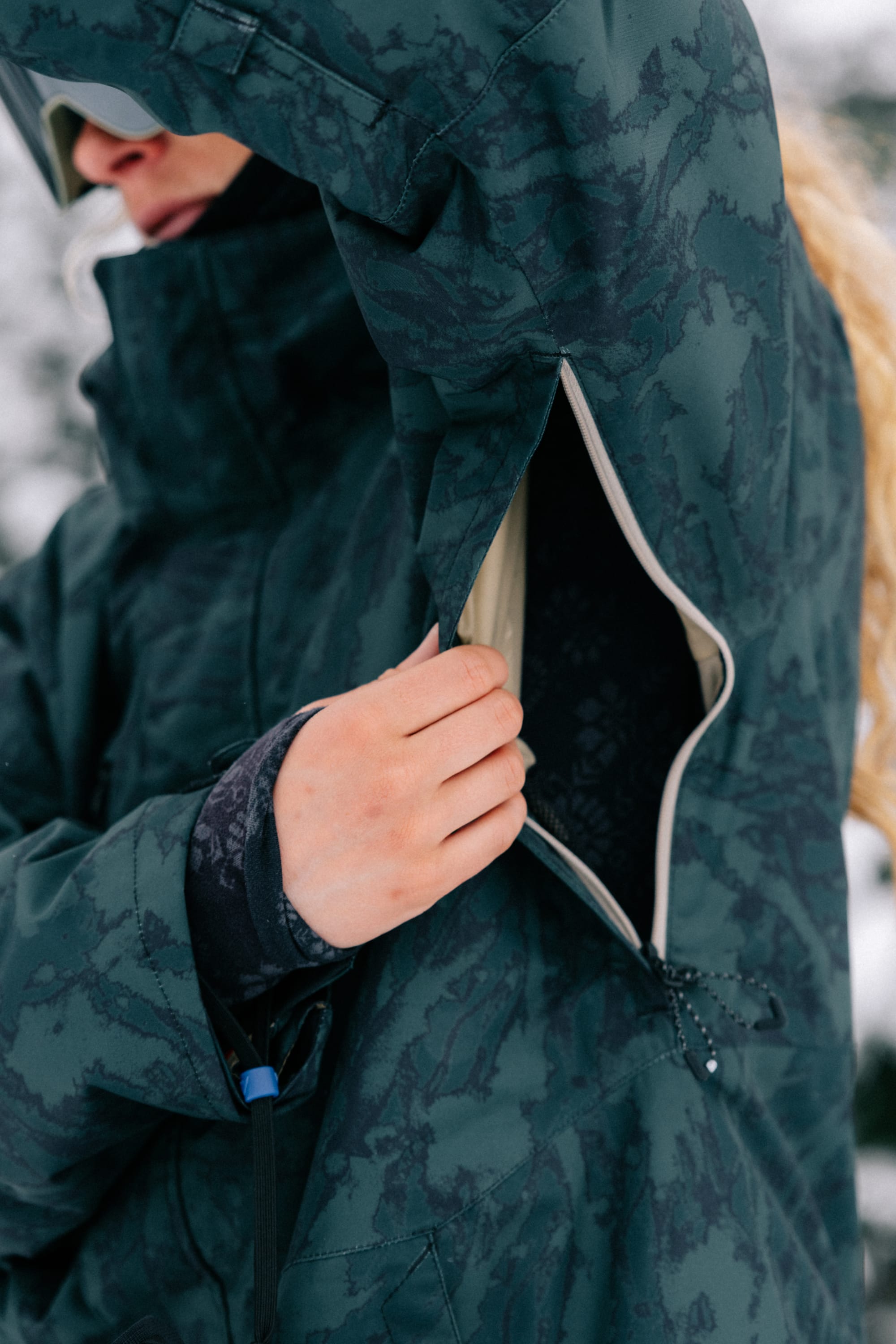 A person wearing the 686 Women’s GORE-TEX® Willow™ Insulated Jacket unzips a vent under their arm. Their hand is visible opening the zipper, with part of their face partially out of frame against a blurred snowy background.