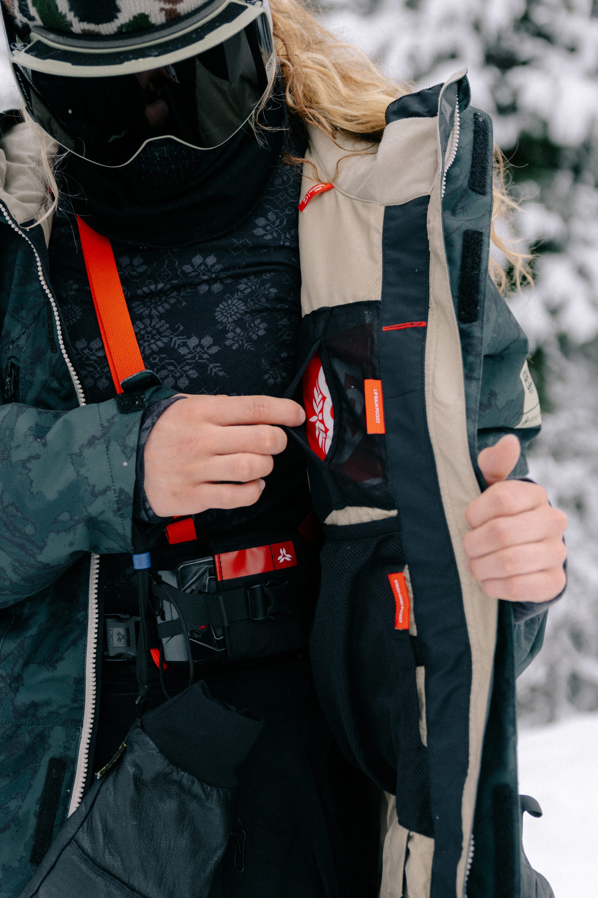 A person dressed for winter opens their 686 Women’s GORE-TEX® Willow™ Insulated Jacket, revealing a red and white patch inside, with snowy trees in the background.