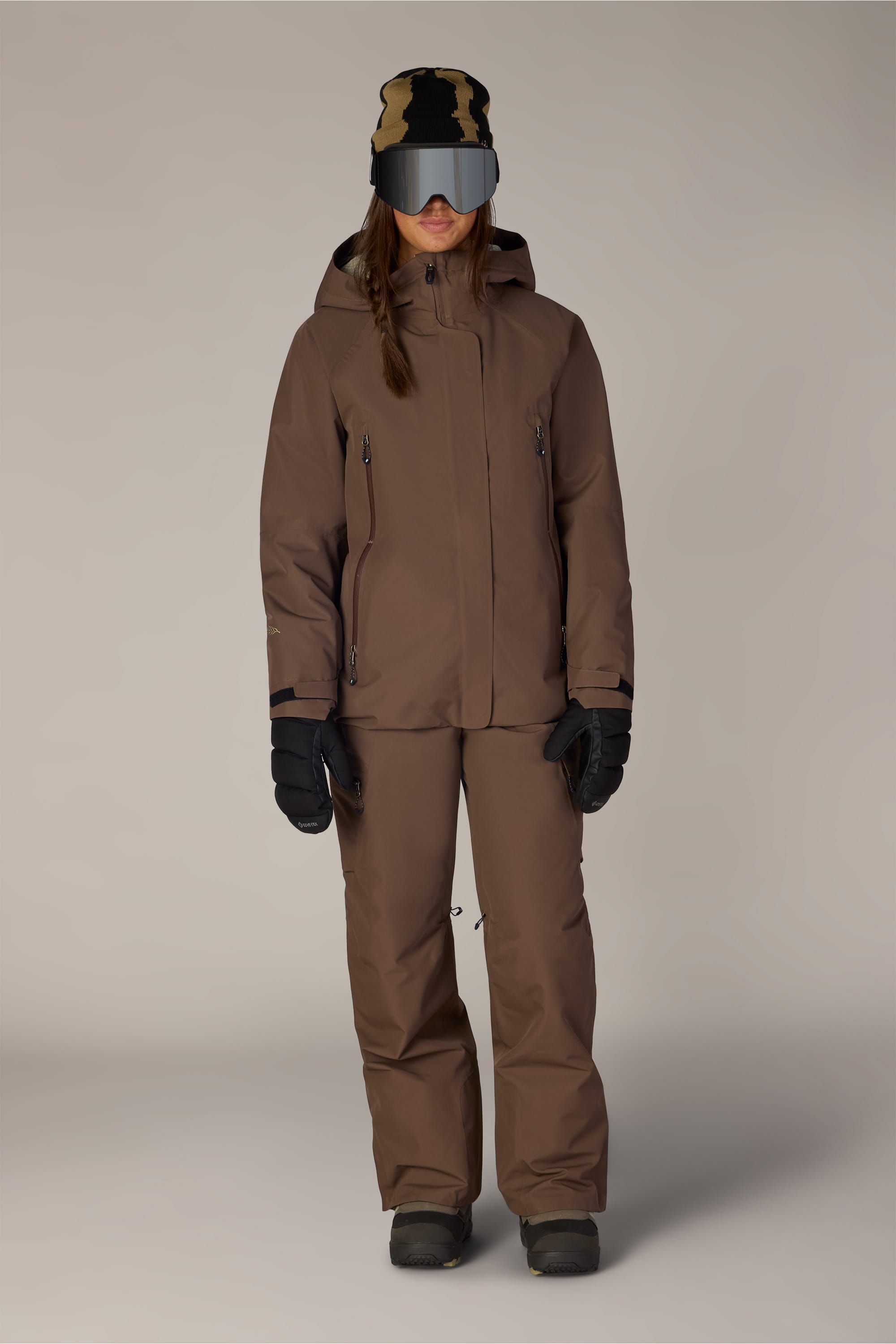 A person wearing the 686 Women’s Whisper™ Insulated Jacket by 686, along with matching pants, black gloves, boots, a patterned beanie, and mirrored ski goggles stands facing forward against a plain background.