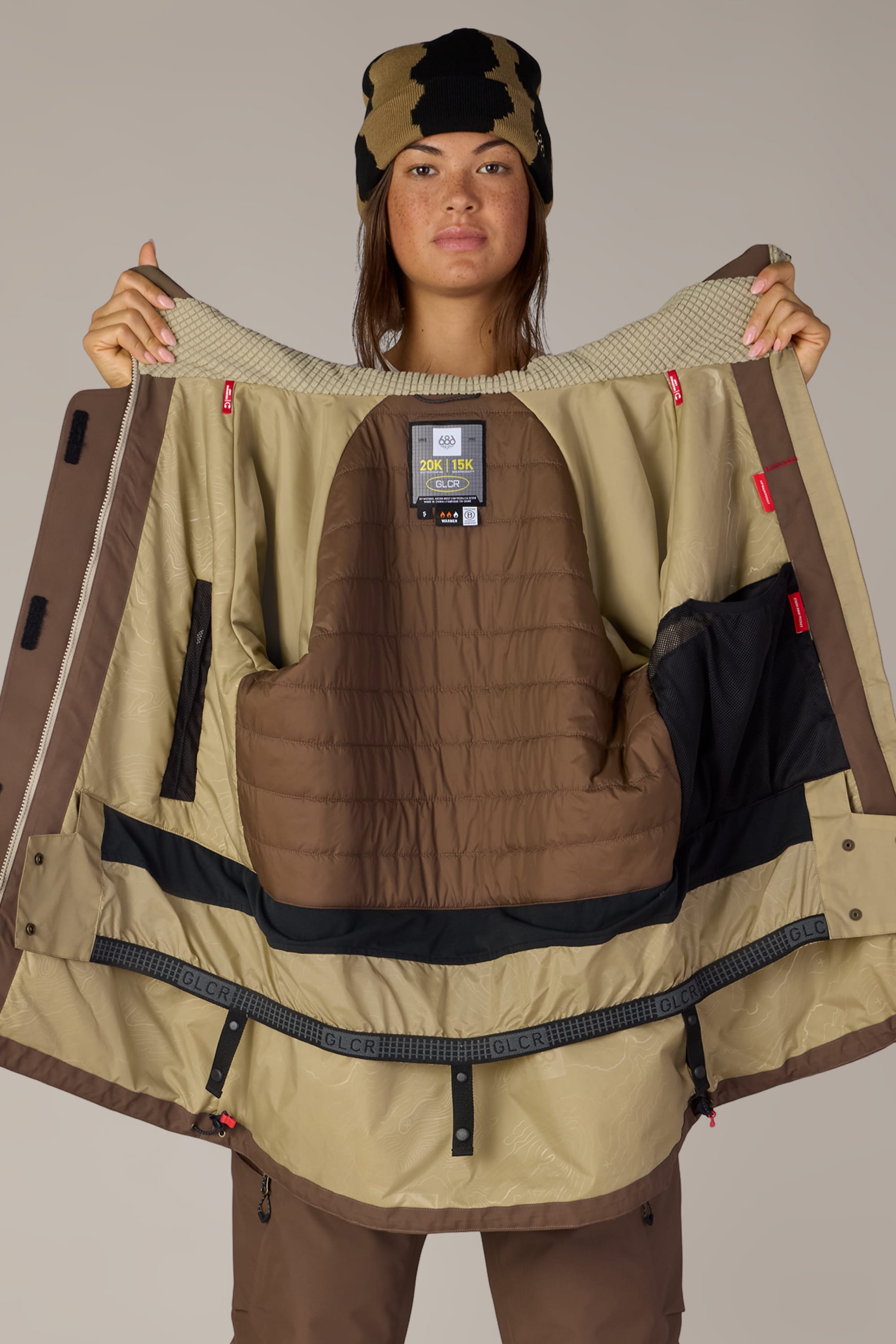 A person wearing brown winter clothing and a camouflage beanie holds open the 686 Women’s Whisper™ Insulated Jacket by 686, displaying its tan and brown interior lining and label while facing the camera against a plain background.