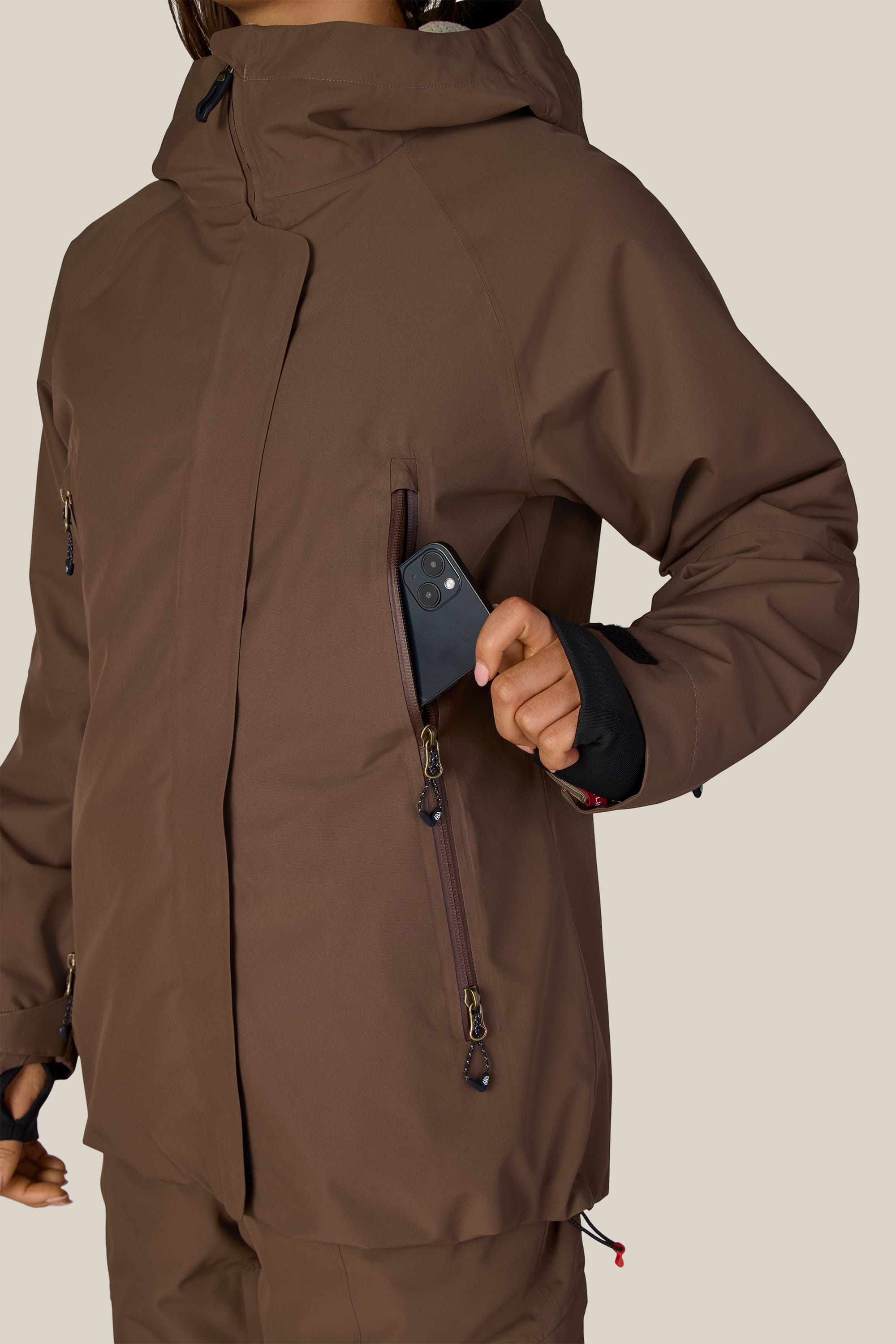 A person wearing the 686 Women’s Whisper™ Insulated Jacket in brown is placing a black smartphone into the jacket’s chest pocket. The 686 jacket features several zippered pockets, and the background is plain.