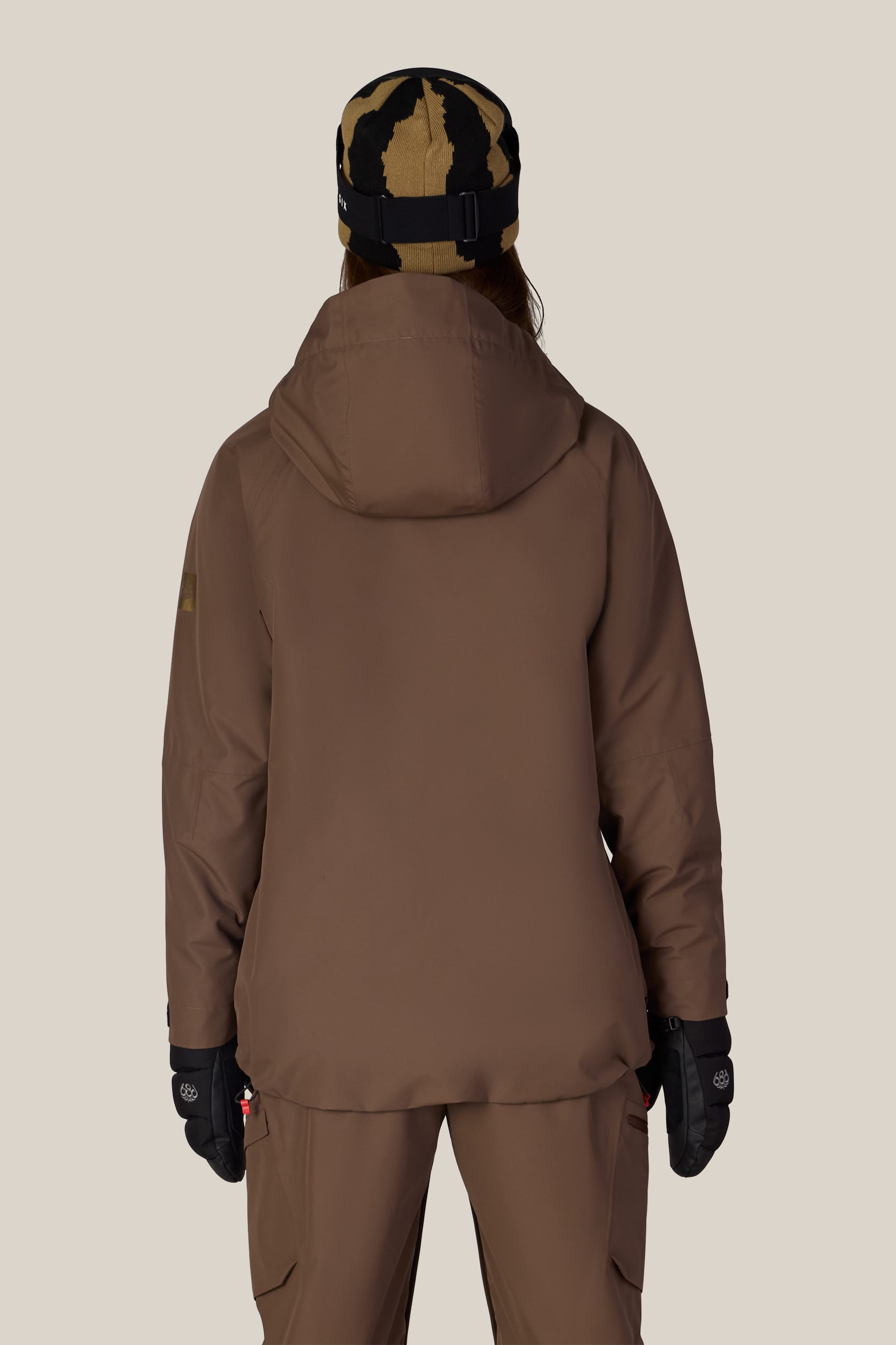 A person is shown from the back against a plain background wearing the 686 Women’s Whisper™ Insulated Jacket by 686, along with brown pants, black gloves, and a tan and black patterned beanie.