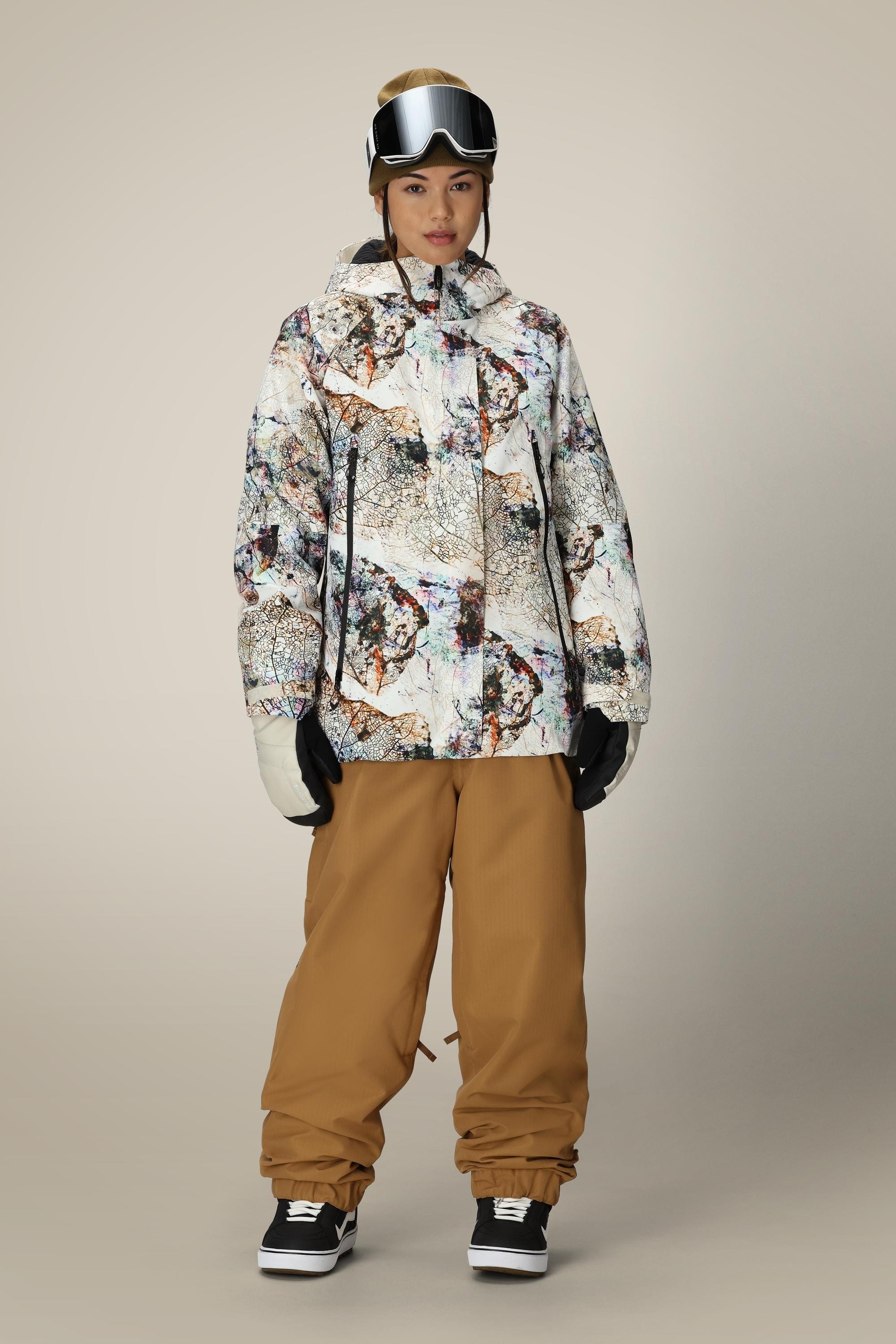 A person stands facing forward, wearing the 686 Womens Whisper Insulated Jacket with a patterned design, loose tan snow pants, black gloves, white mittens, black snow boots, ski goggles, and a beige beanie against a neutral background.