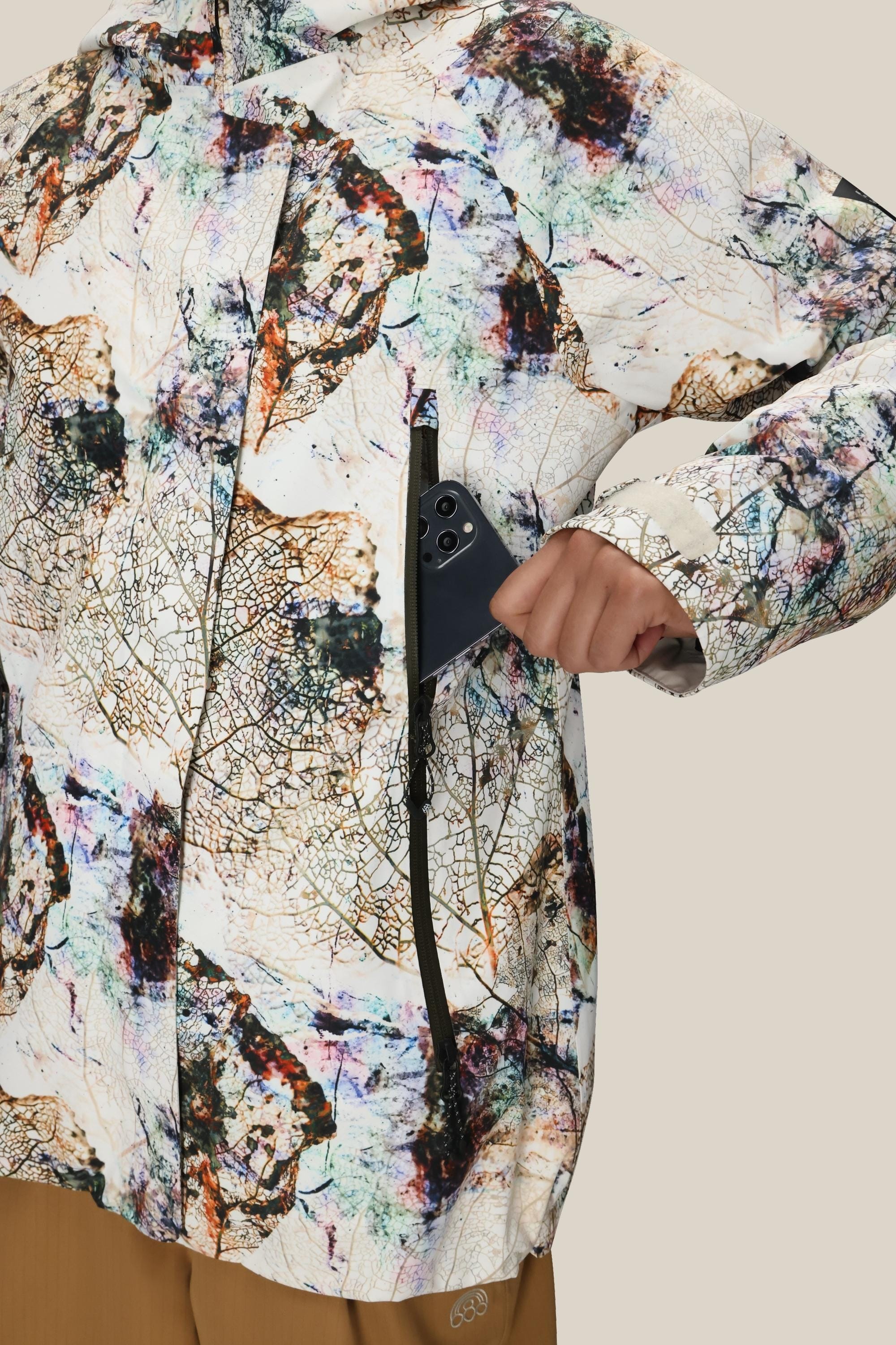 A person wearing the 686 Womens Whisper Insulated Jacket with a colorful abstract pattern places a black smartphone into its zippered chest pocket. The beige background and non-visible face keep the focus on the jacket.
