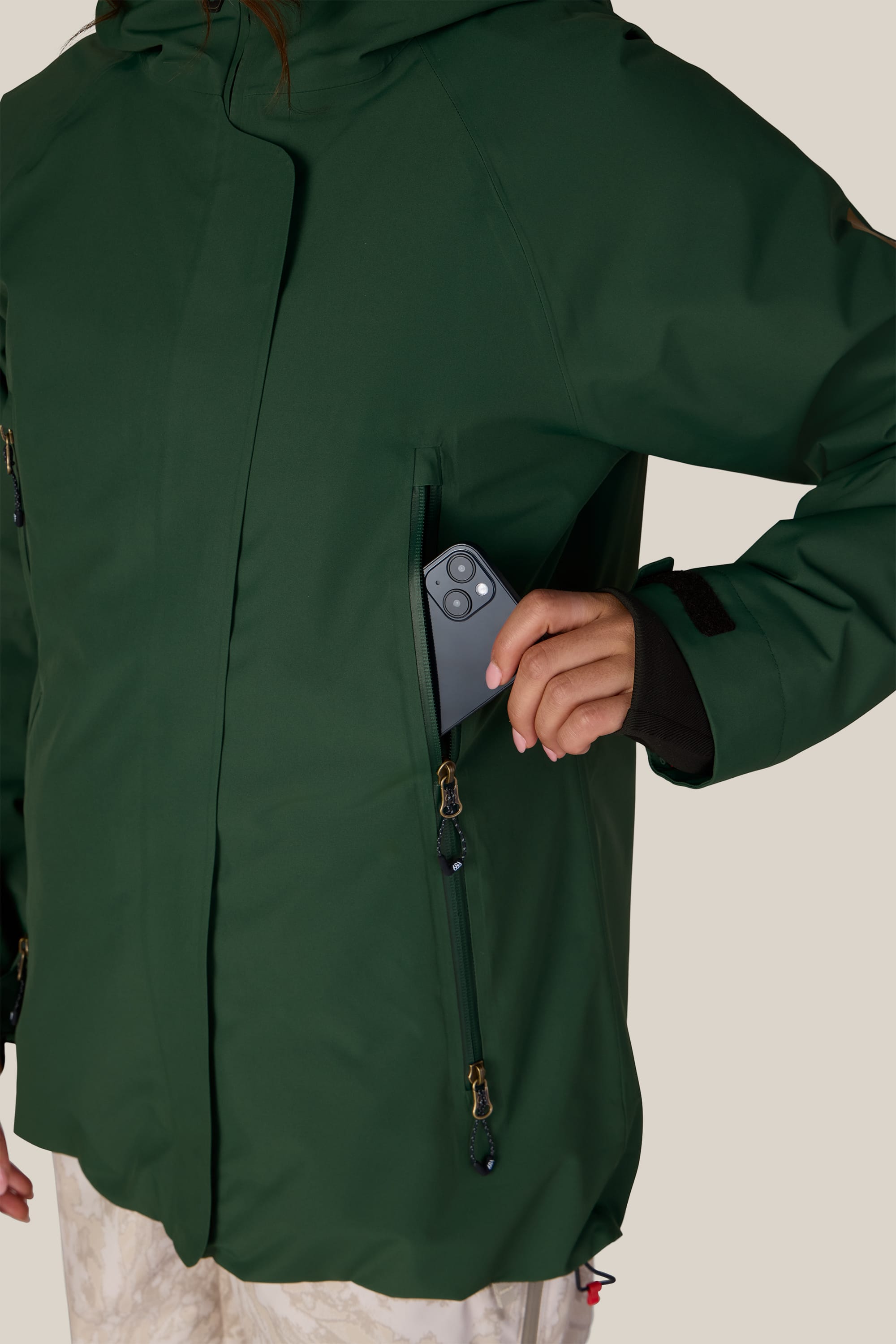 A person in a 686 Women’s Whisper™ Insulated Jacket slips a smartphone into the jacket’s zippered chest pocket, with only their hand and part of the phone visible.