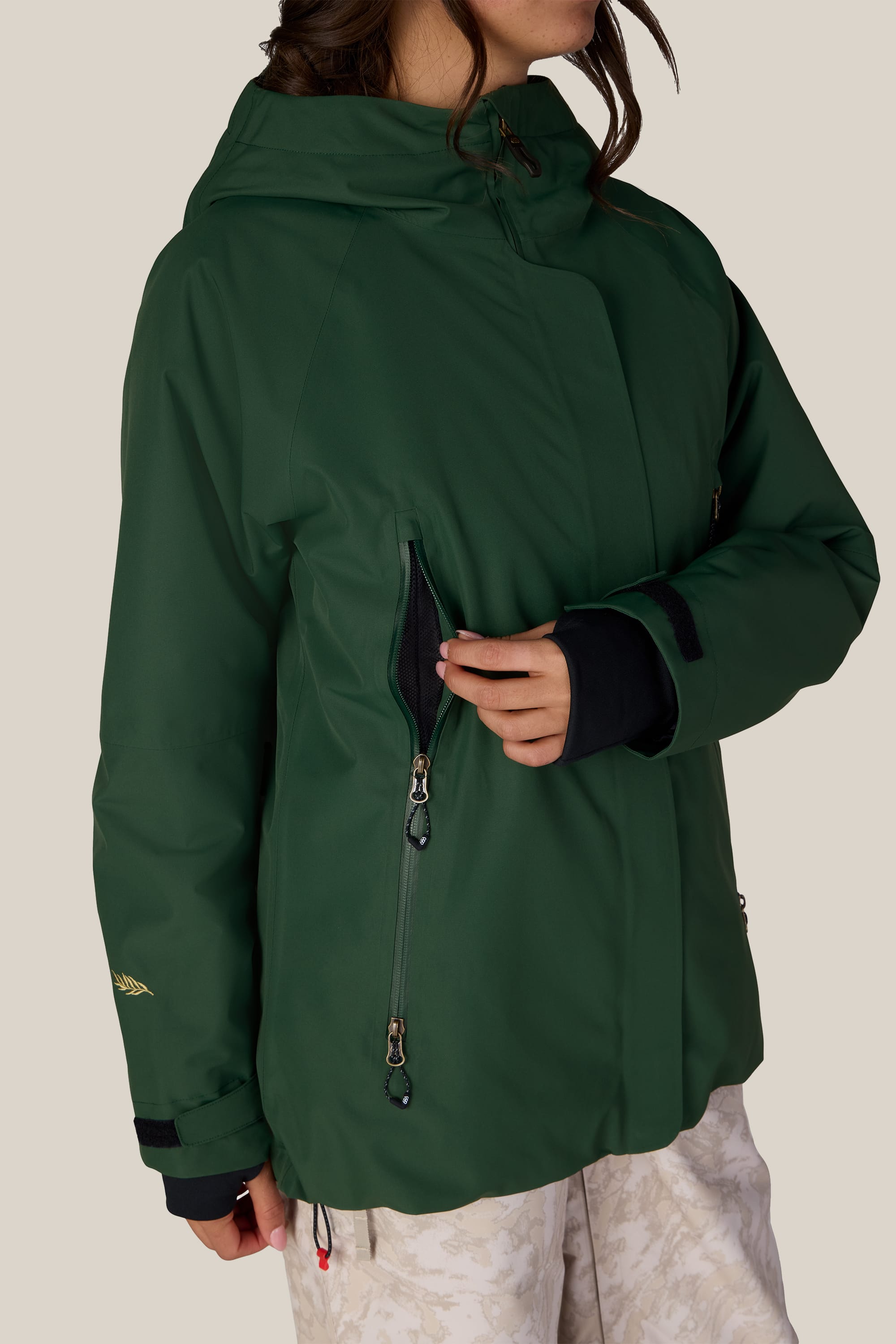 A person wearing the 686 Women’s Whisper™ Insulated Jacket in green unzips a chest pocket, their face partially out of frame, against a plain background.