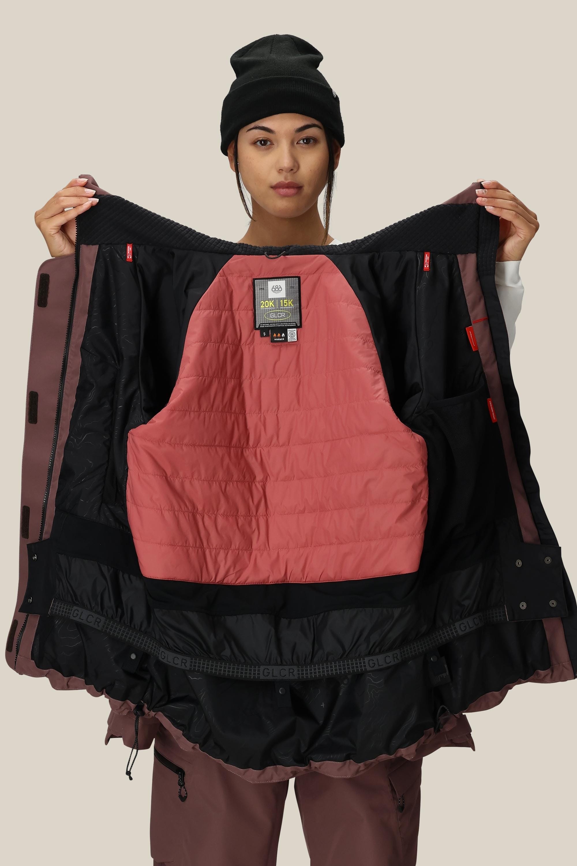 A woman in a black beanie and mauve snow pants showcases the 686 Womens Whisper Insulated Jacket in mauve and black, revealing its padded pink interior and multiple pockets, standing against a plain beige background.