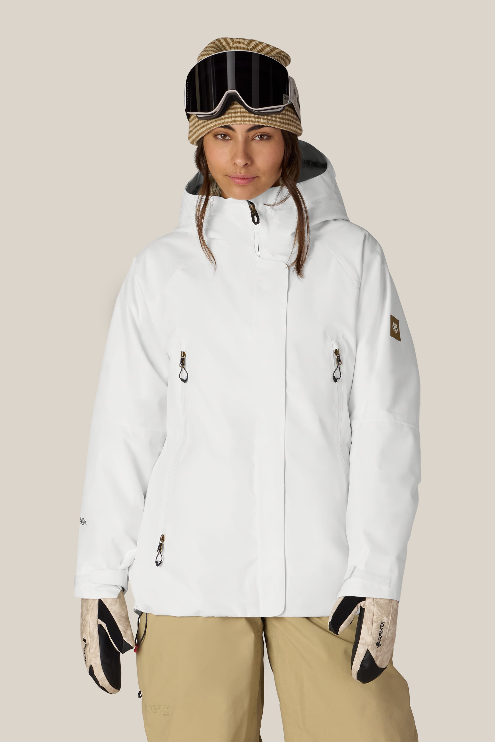 A woman wearing the 686 Women’s Whisper™ Insulated Jacket stands facing the camera against a plain background, paired with tan snow pants, gloves, and ski goggles.