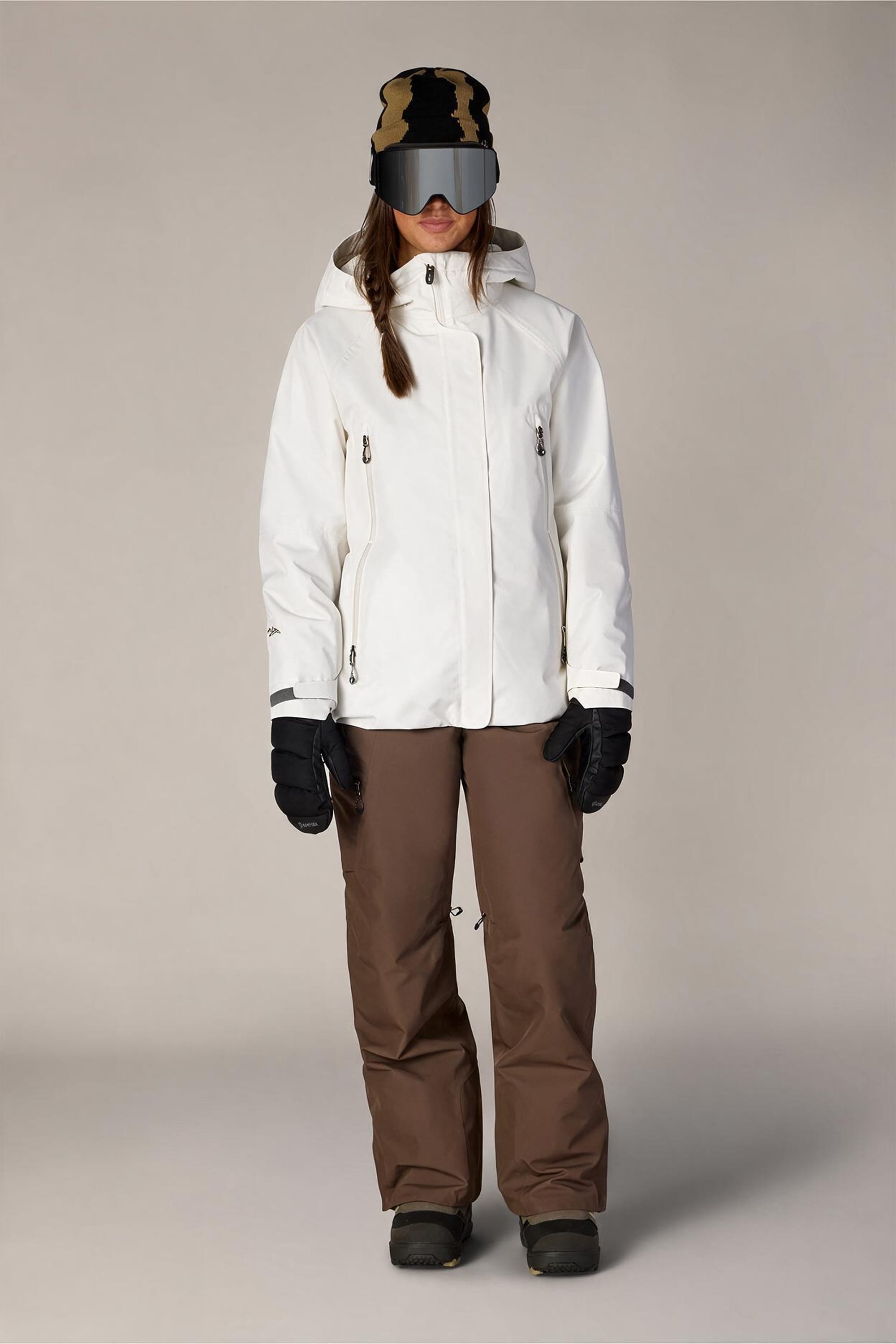 A person wearing the 686 Women’s Whisper™ Insulated Jacket by 686, paired with brown snow pants, black gloves, snow boots, a camouflage hat, and large reflective ski goggles, standing against a plain light background.