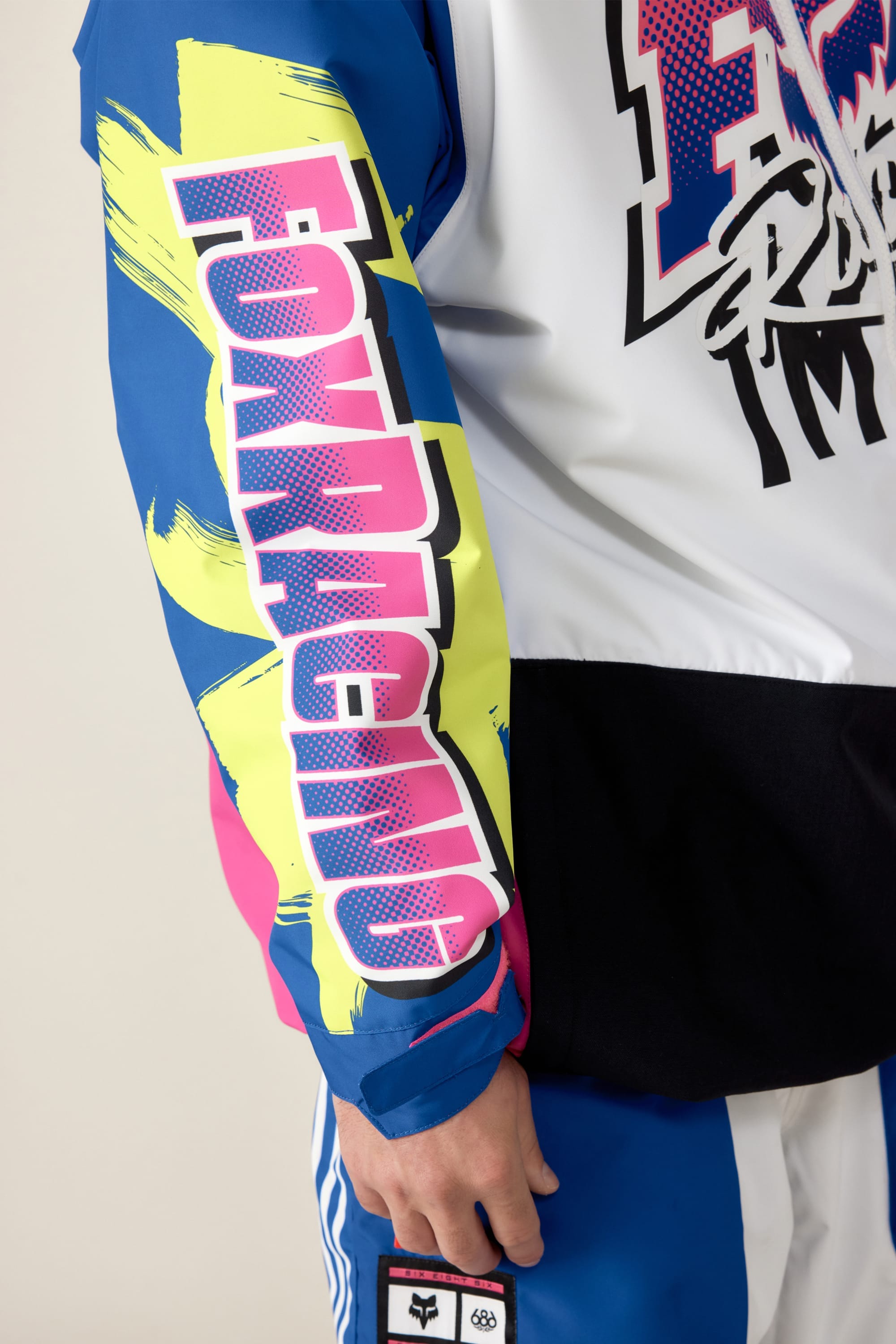 A close-up of a person wearing the 686 Men's Fox Racing Shell Anorak by 686, featuring a blue sleeve with yellow and pink graphics, bold FOX RACING text, and black, white, and blue color sections.