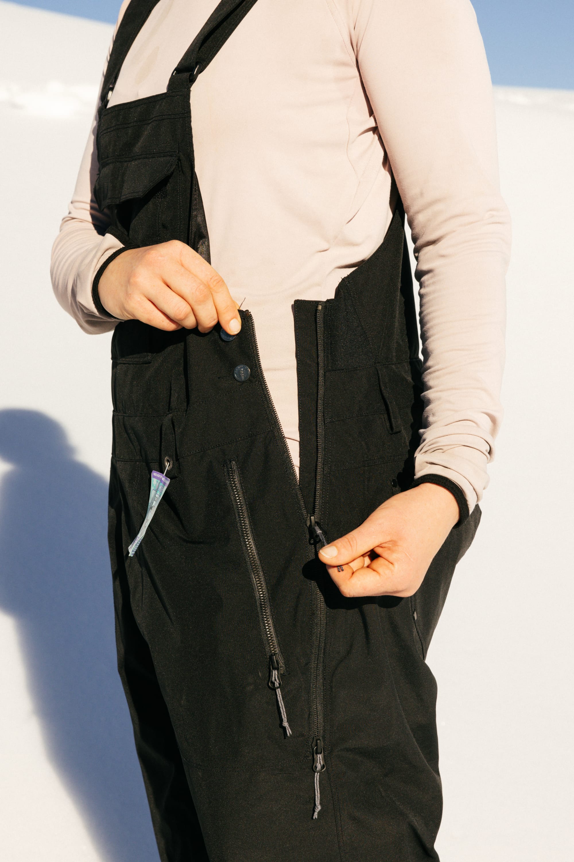 A person wearing a light long-sleeve shirt and the 686 Women’s Black Magic™ Insulated Bib is unzipping a side zipper on the bib while standing on snow in bright sunlight. Only their torso and hands are visible.