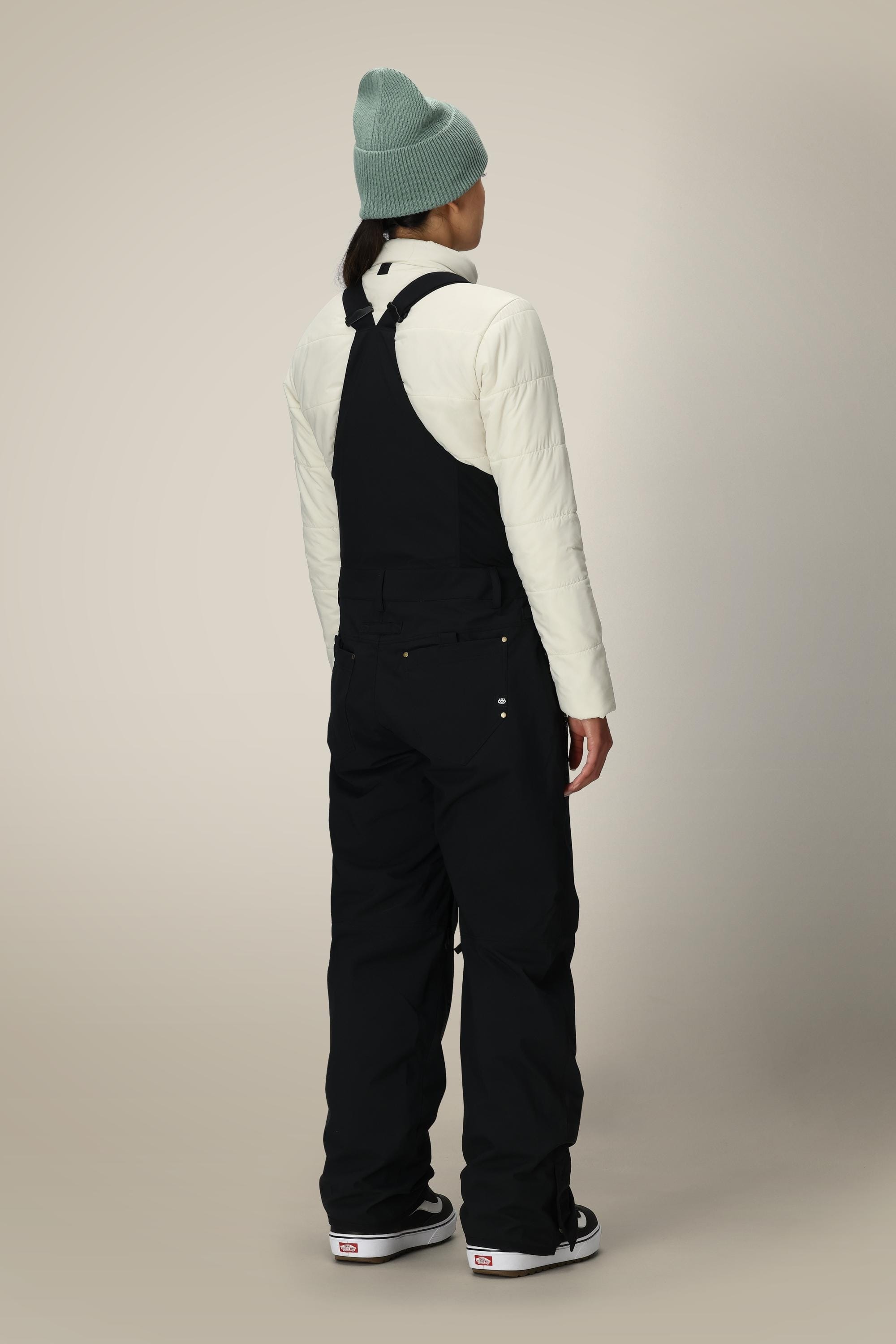 A person is seen from behind wearing the 686 Women’s Black Magic™ Insulated Bib by 686, paired with a light-colored jacket, black shoes, and a light green beanie against a plain background.
