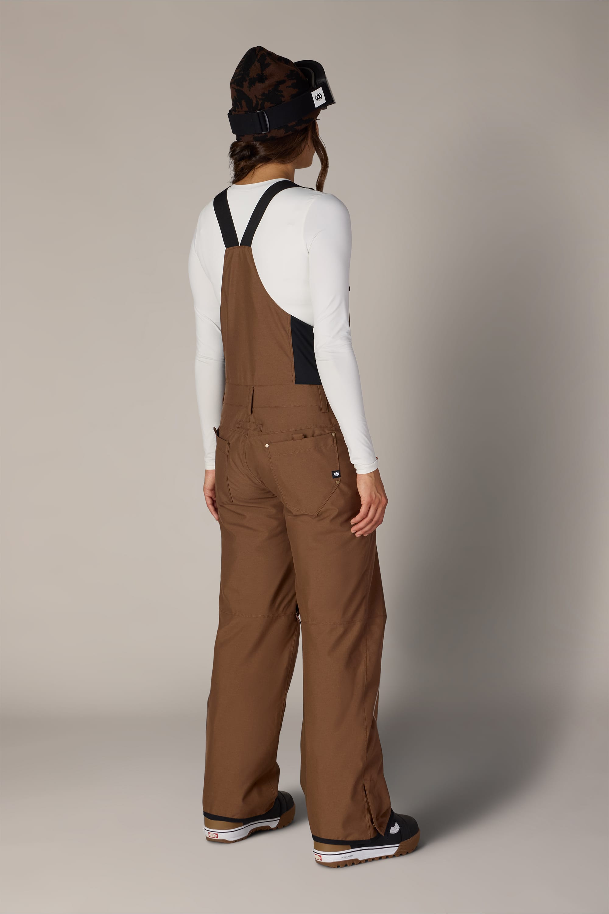A person stands facing away, wearing the 686 Women’s Black Magic™ Insulated Bib over a white long-sleeve shirt, paired with a black beanie and black shoes. The background is plain and light gray.