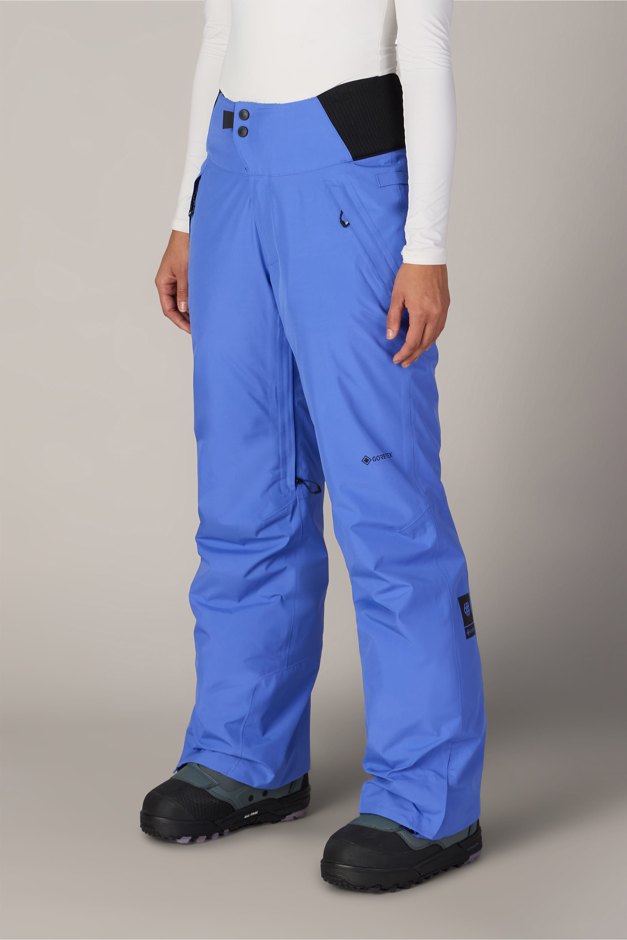 A person is wearing the 686 Women’s GORE-TEX® Willow™ Insulated Pant in a bright blue color, paired with black snow boots and a long-sleeved white top, standing against a plain background.