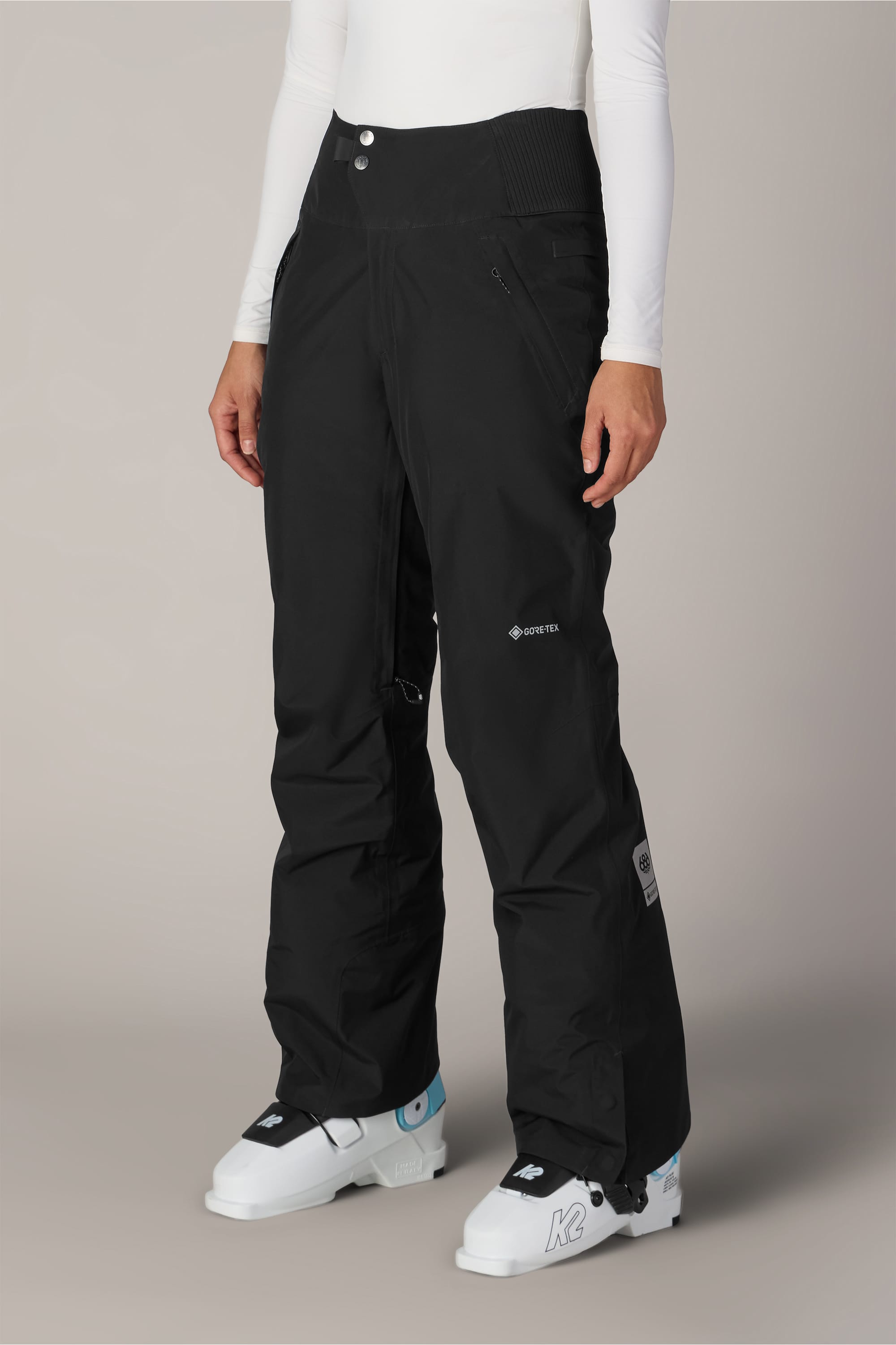 A person wearing the 686 Women’s GORE-TEX® Willow™ Insulated Pant in black, paired with a white long-sleeve top and white ski boots, standing against a plain light background.