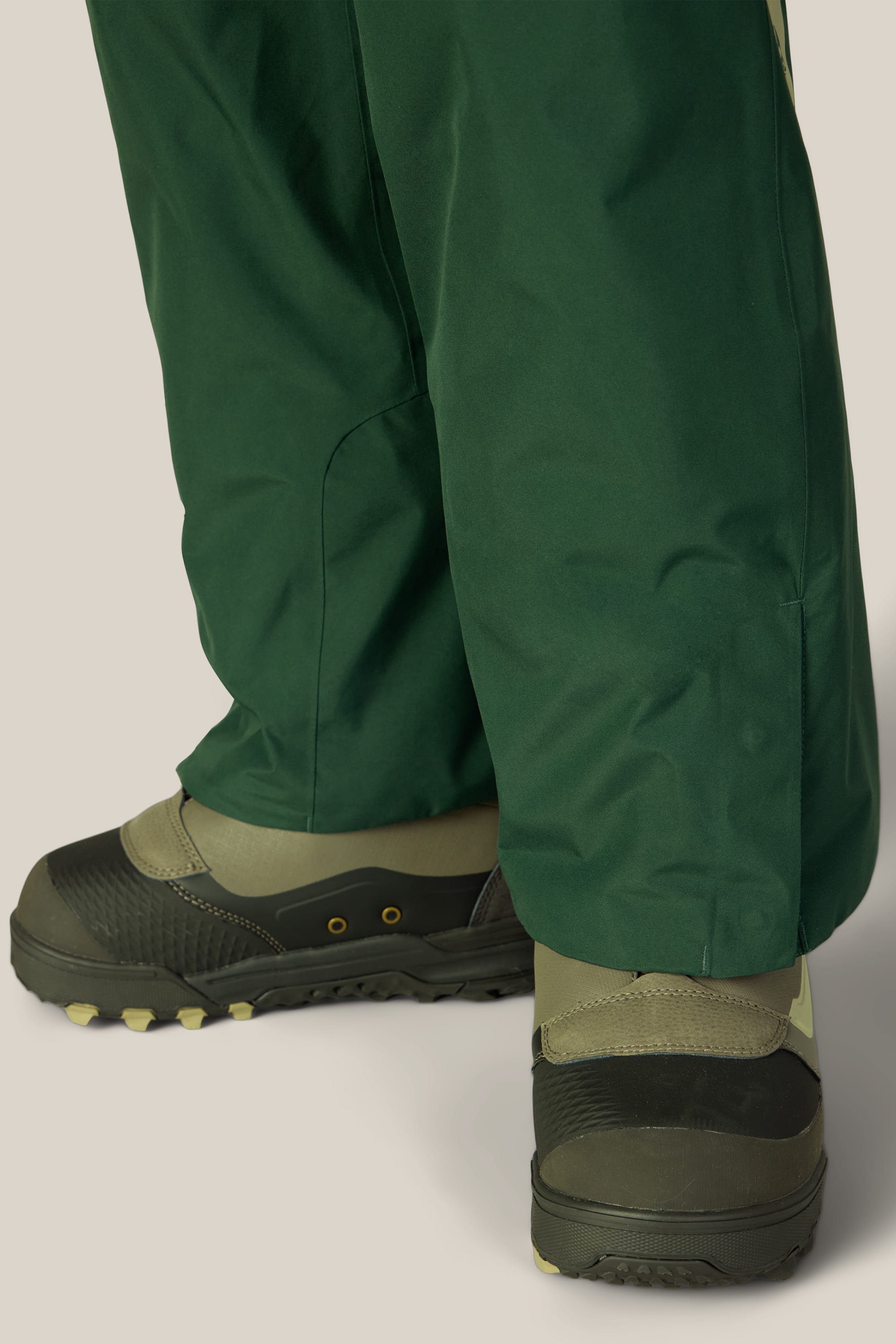 A close-up of a person wearing the 686 Women’s GORE-TEX® Willow™ Insulated Pant and dark green outdoor shoes with thick soles, standing on a light background.