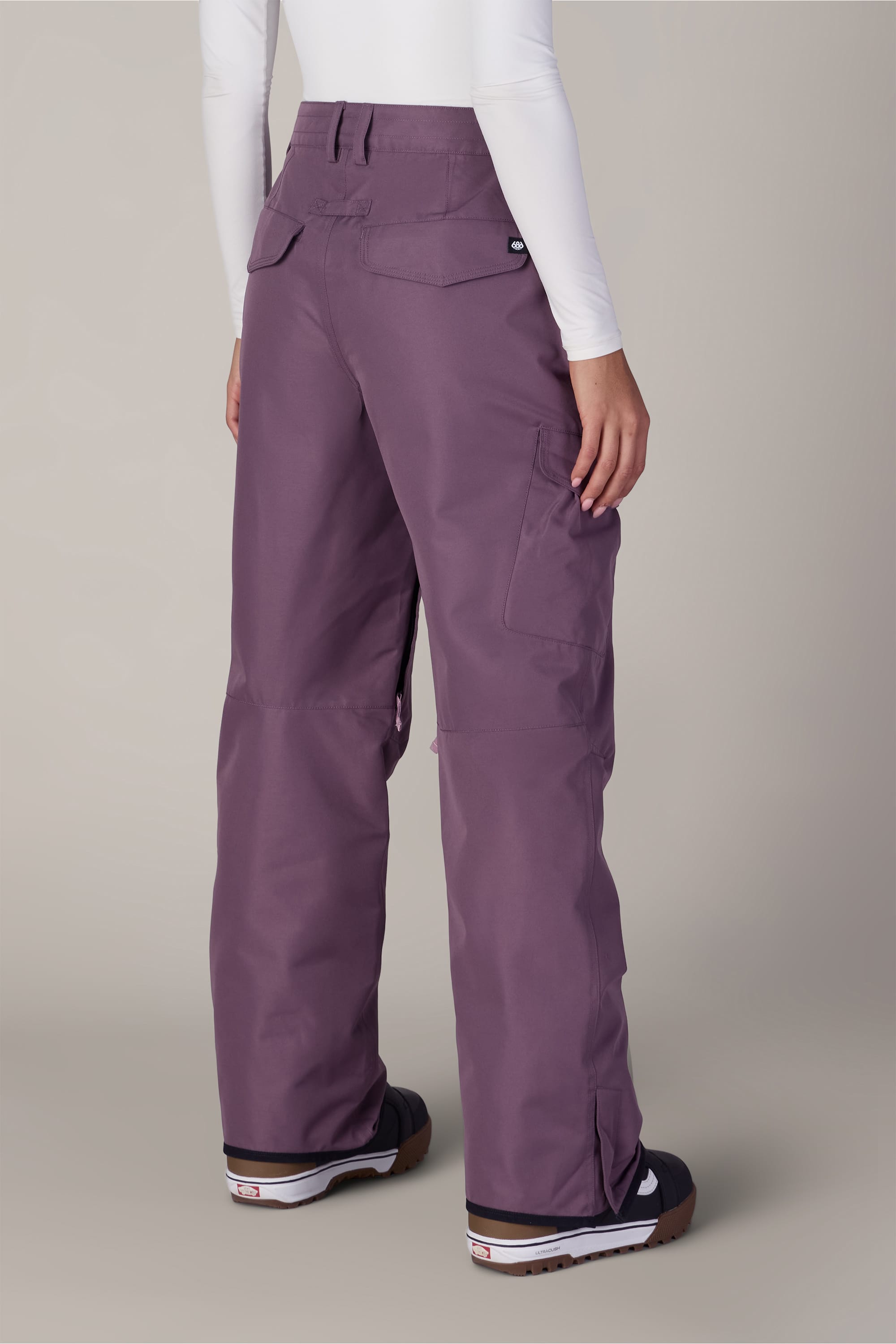A person wearing the 686 Women’s Aura™ Insulated Cargo Pant in purple and black shoes stands with their back to the camera, also sporting a white long-sleeve top.