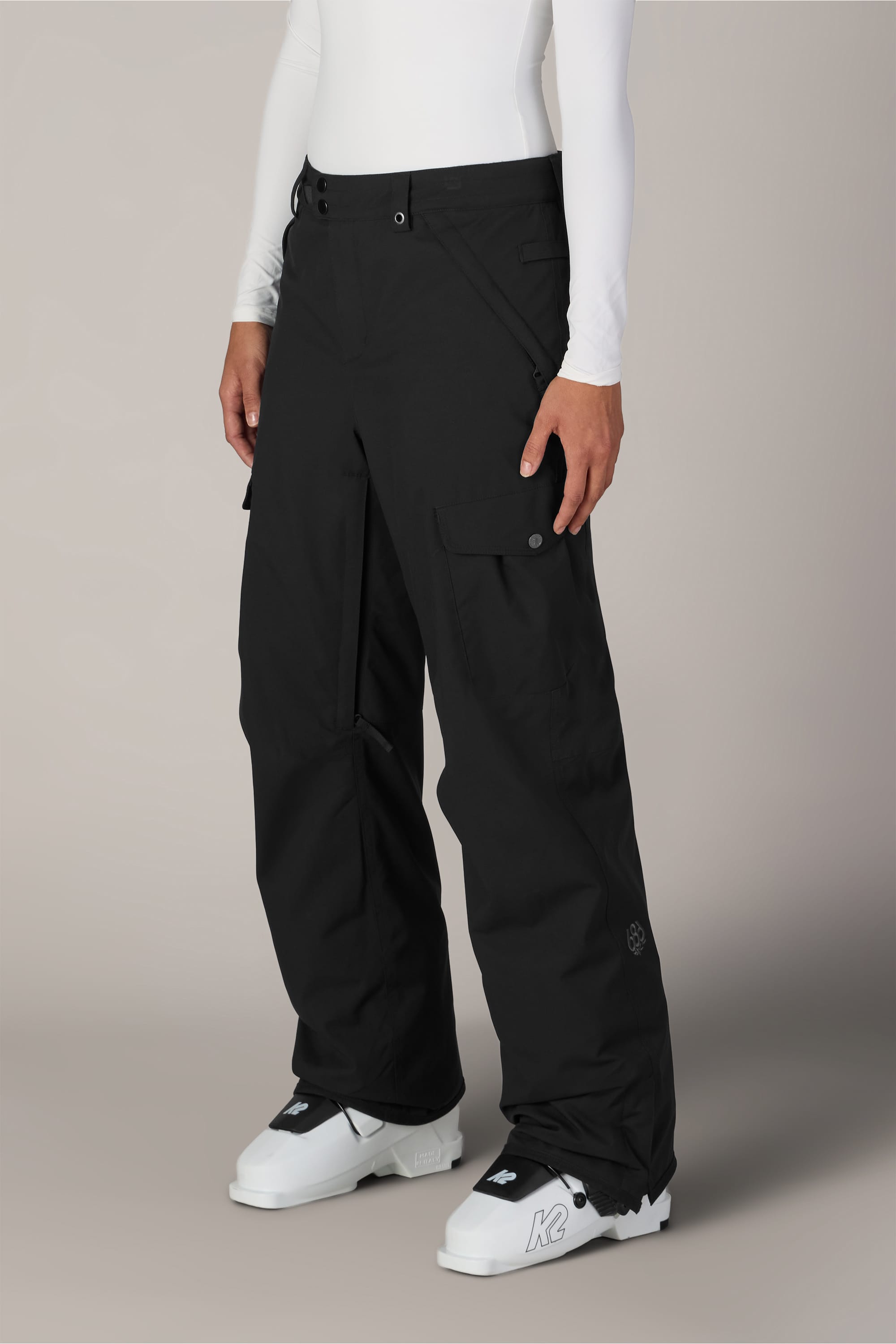 A person wearing the 686 Women’s Aura™ Insulated Cargo Pant in black, a white long-sleeve top, and white snow boots stands against a neutral background.