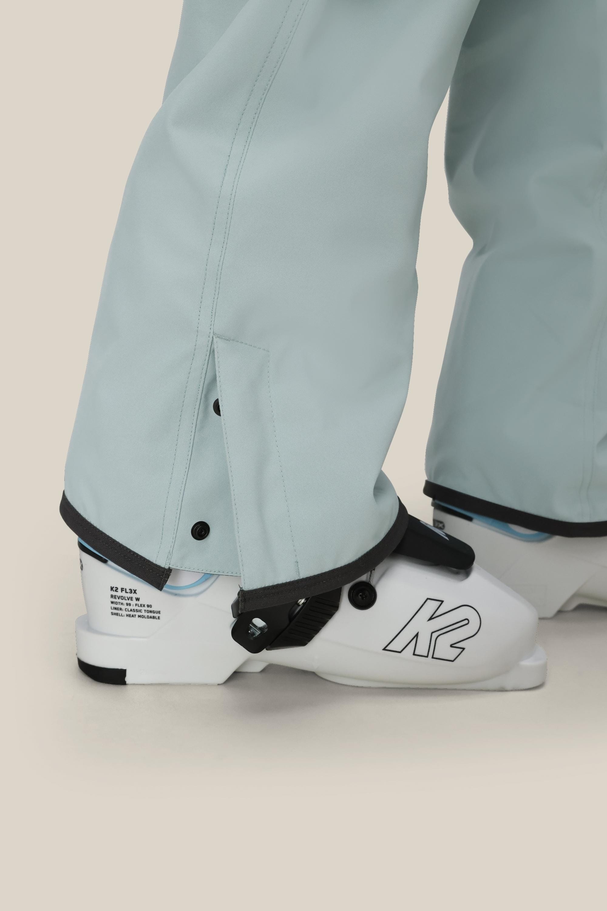 A close-up of a person wearing 686 Womens Aura Insulated Cargo Pant in light blue over white K2 ski boots, with the pant cuffs partially covering the boots.