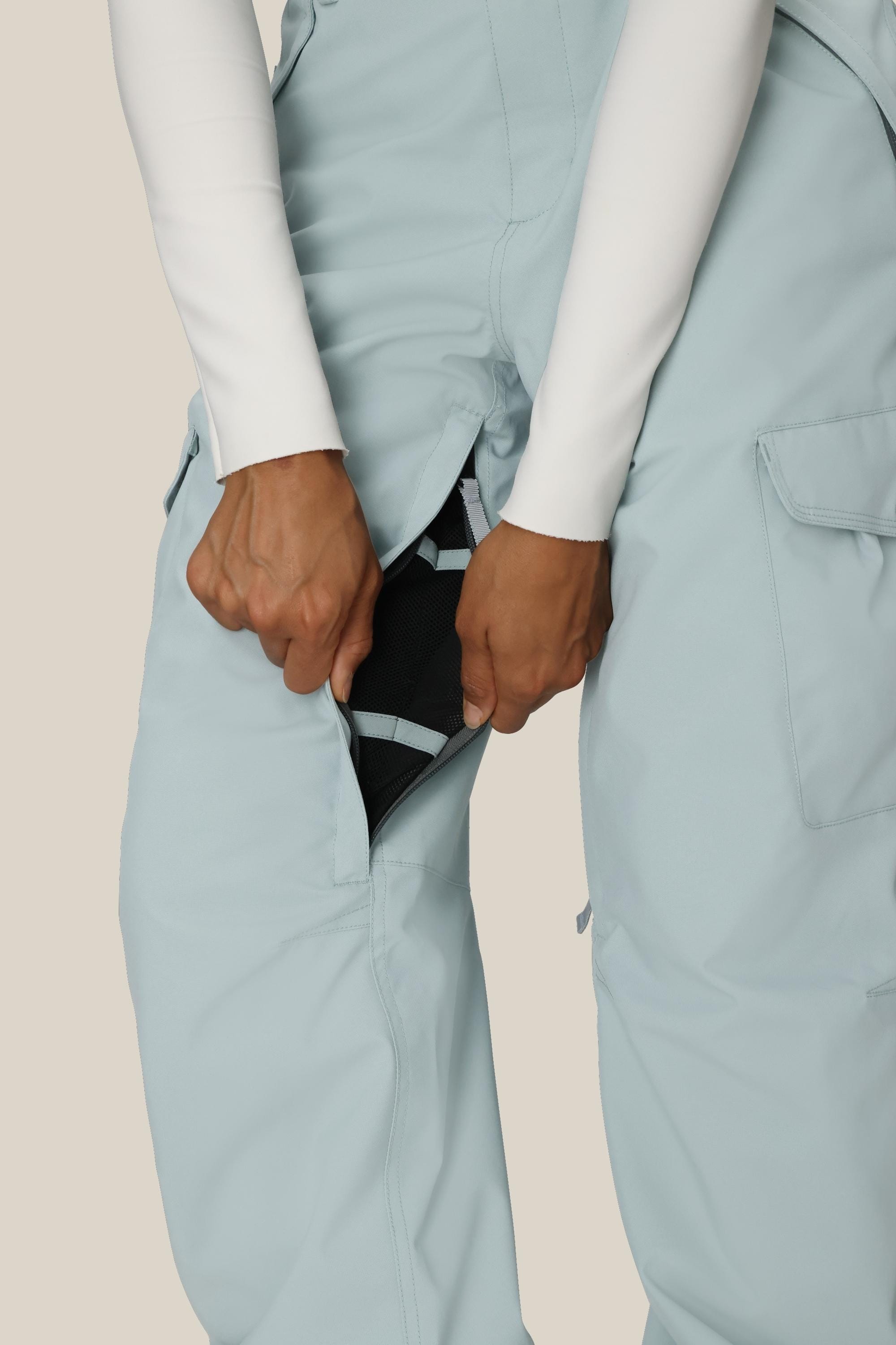A person wearing 686 Womens Aura Insulated Cargo Pant in light blue and a white long-sleeve top unzips a side vent, revealing black mesh fabric underneath.