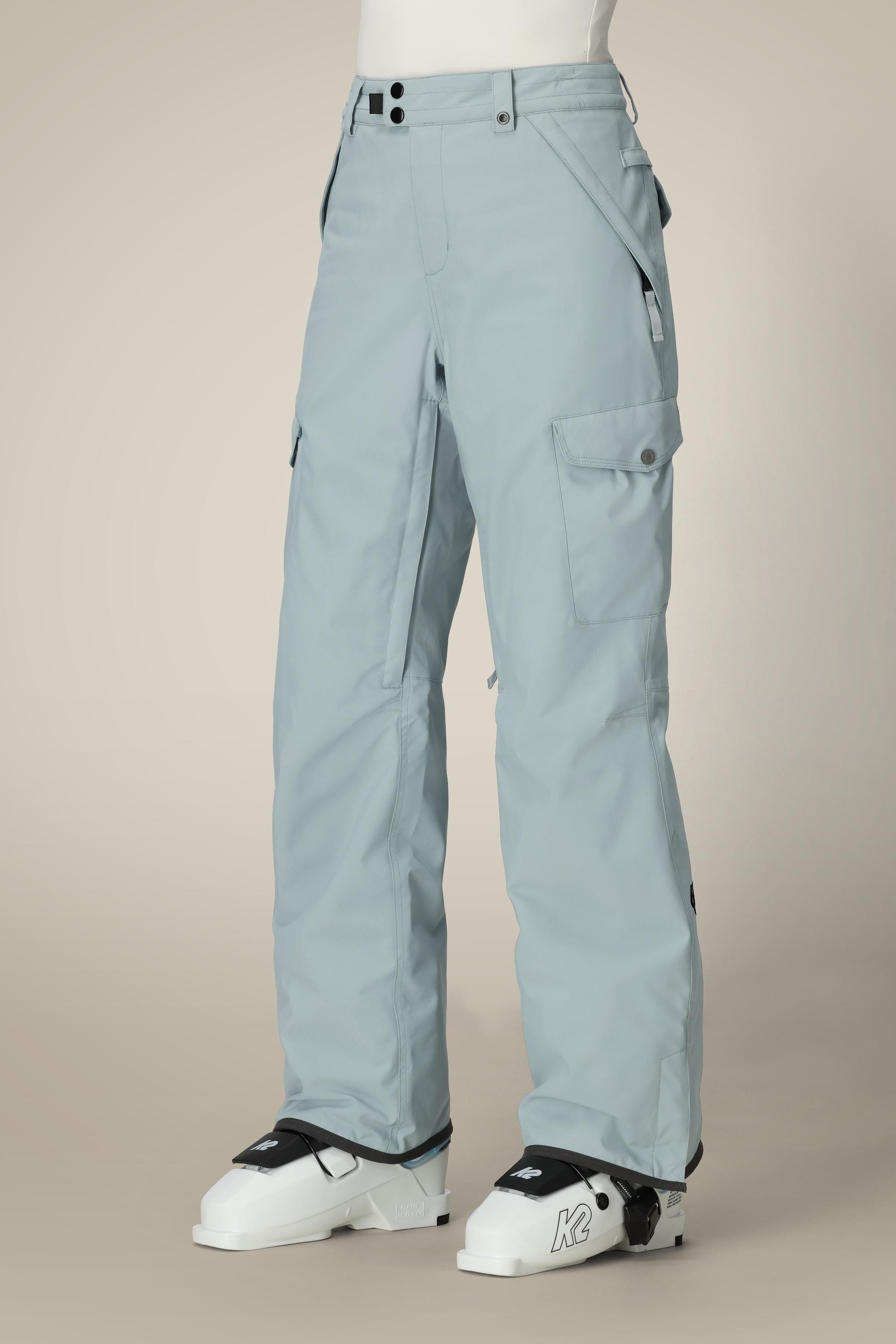A person models 686 Womens Aura Insulated Cargo Pant in light blue with white ski boots, standing against a plain beige background; their upper body is not visible.